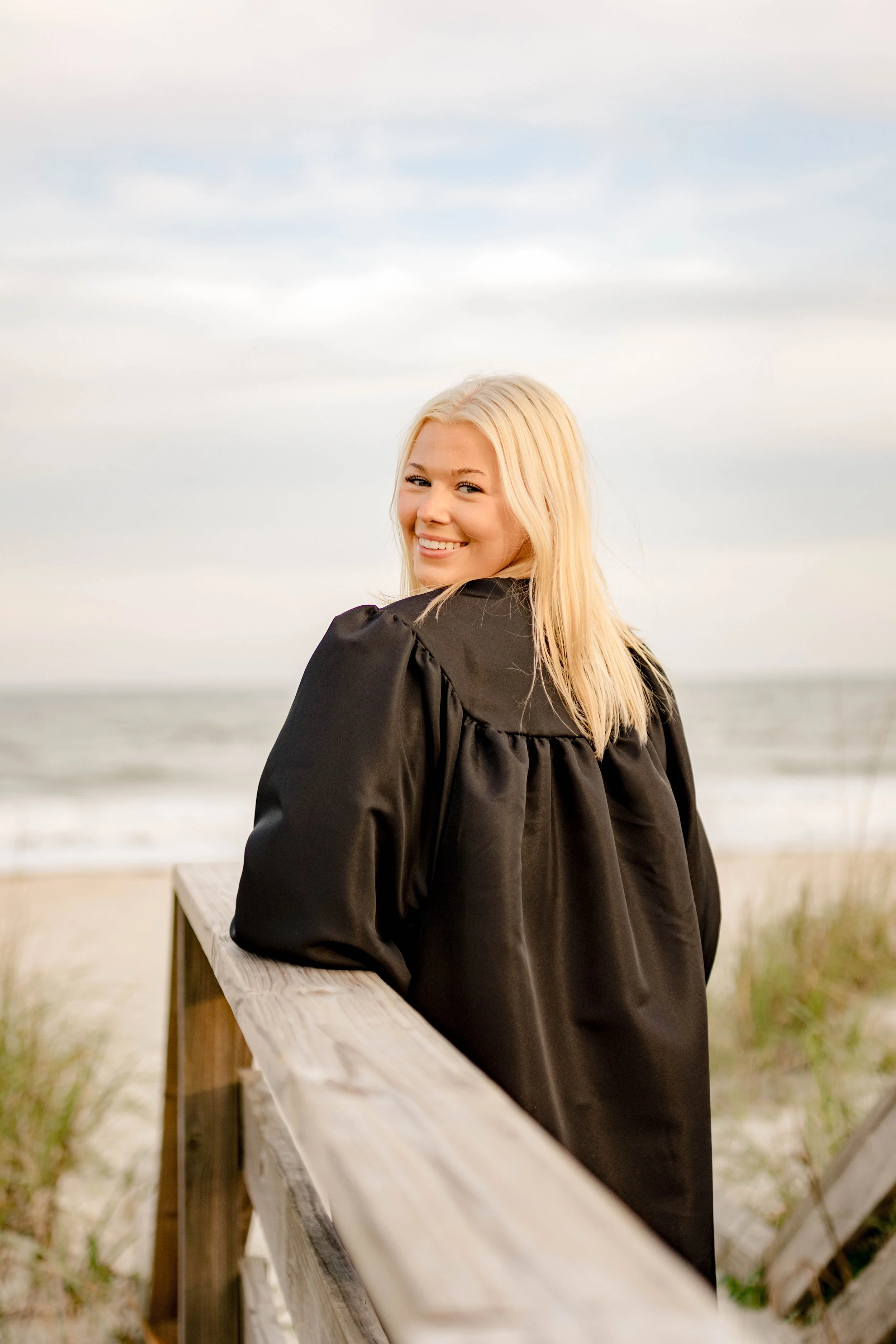 High school senior in Myrtle Beach wearing cap and gown during golden hour beach photo session, splashing in the water and smiling, taken by Myrtle Beach family and senior photographer.