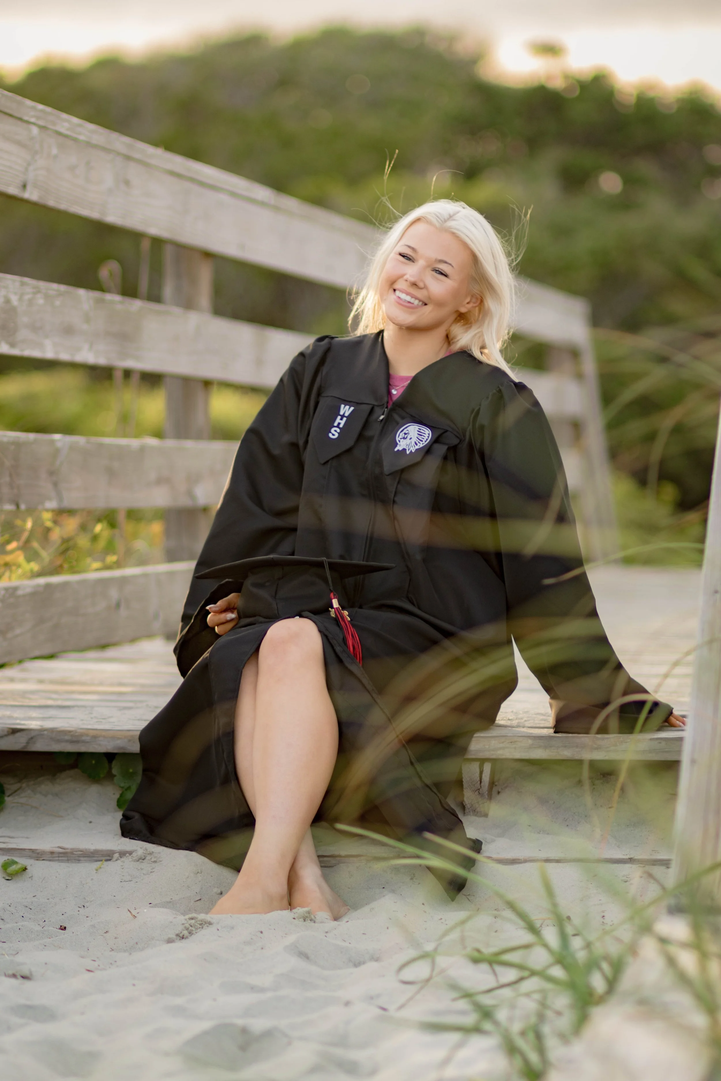 High school senior in Myrtle Beach wearing cap and gown during golden hour beach photo session, splashing in the water and smiling, taken by Myrtle Beach family and senior photographer.
