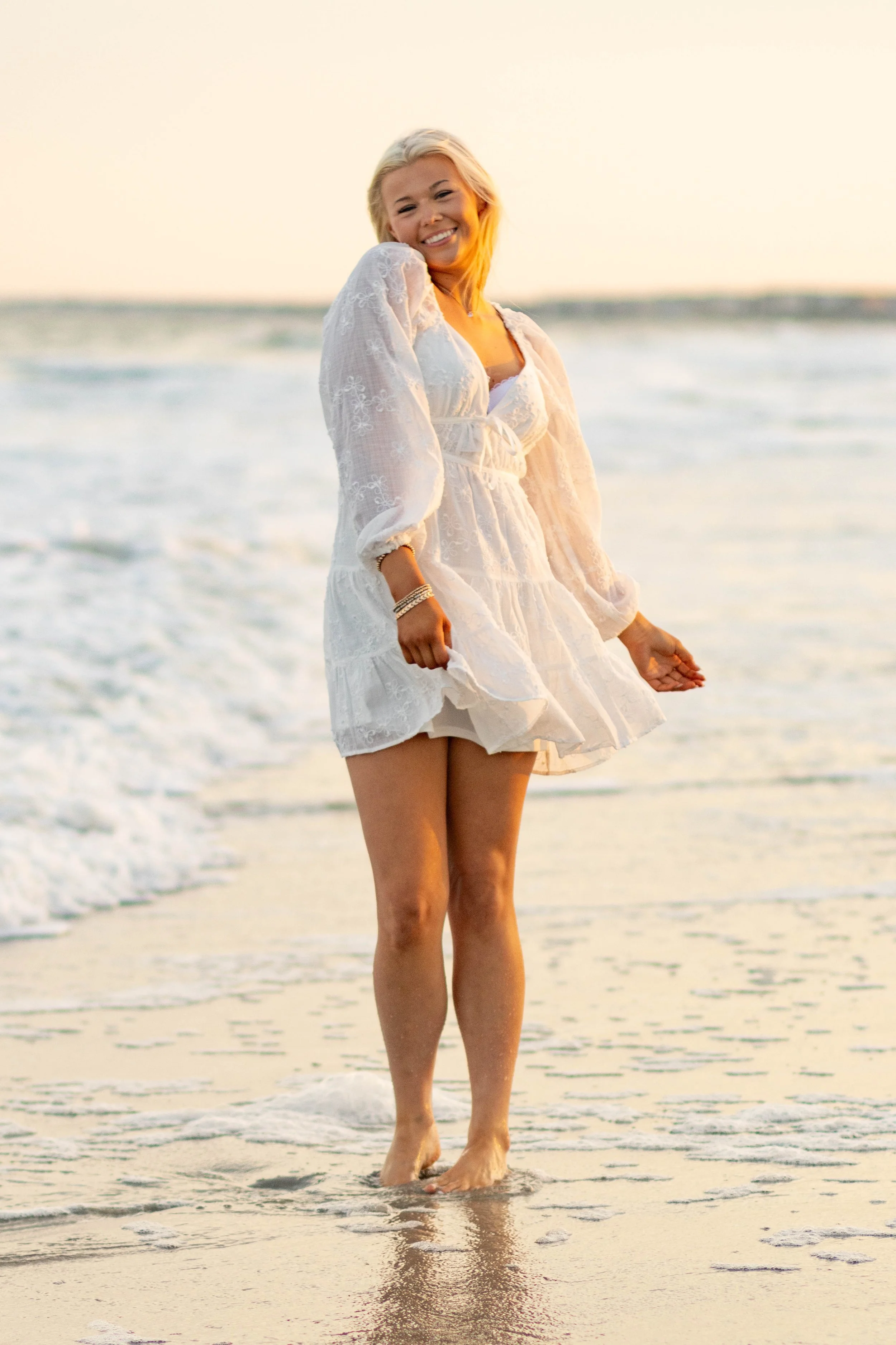 High school senior in Myrtle Beach wearing cap and gown during golden hour beach photo session, splashing in the water and smiling, taken by Myrtle Beach family and senior photographer.