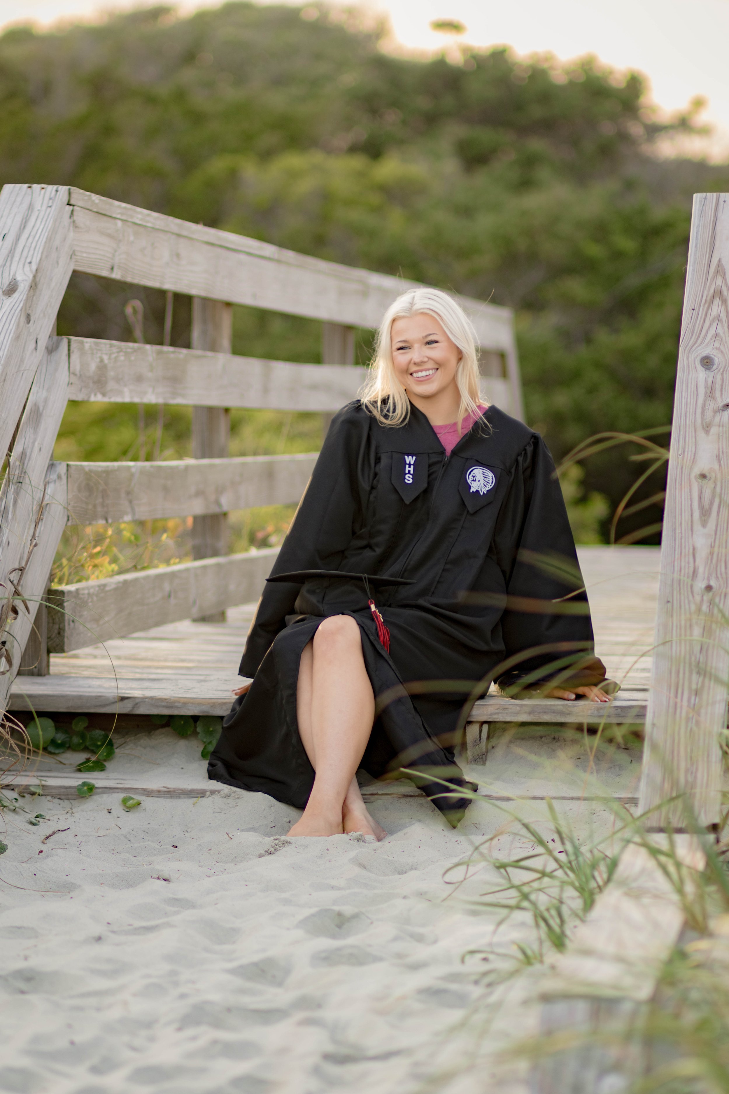 High school senior in Myrtle Beach wearing cap and gown during golden hour beach photo session, splashing in the water and smiling, taken by Myrtle Beach family and senior photographer.