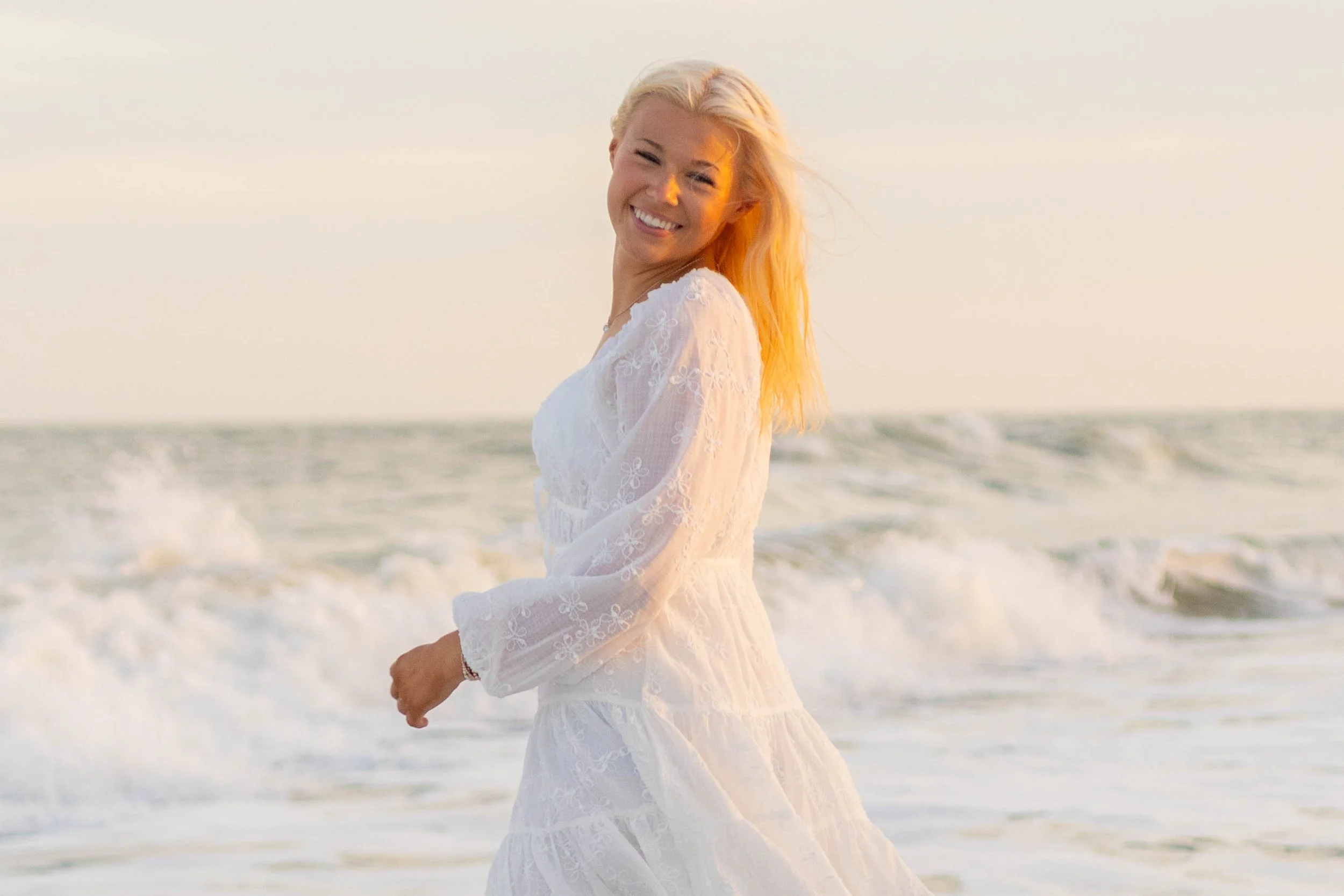 High school senior in Myrtle Beach wearing cap and gown during golden hour beach photo session, splashing in the water and smiling, taken by Myrtle Beach family and senior photographer.