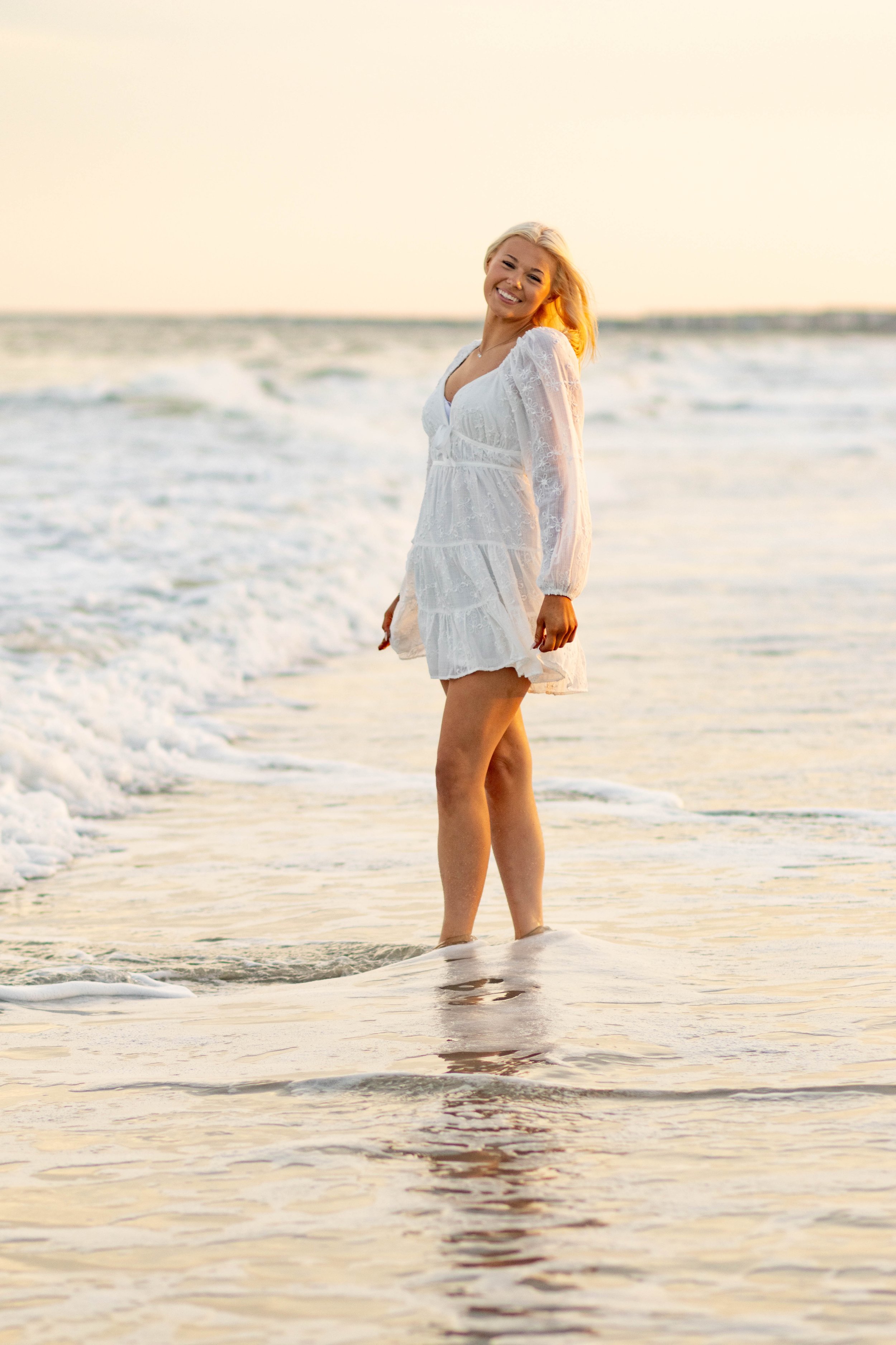 High school senior in Myrtle Beach wearing cap and gown during golden hour beach photo session, splashing in the water and smiling, taken by Myrtle Beach family and senior photographer.