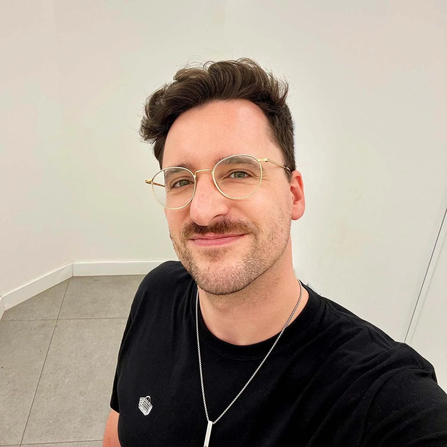 A man with dark, wavy hair, wearing glasses, a black T-shirt, and a necklace, smiling in front of a plain white wall.