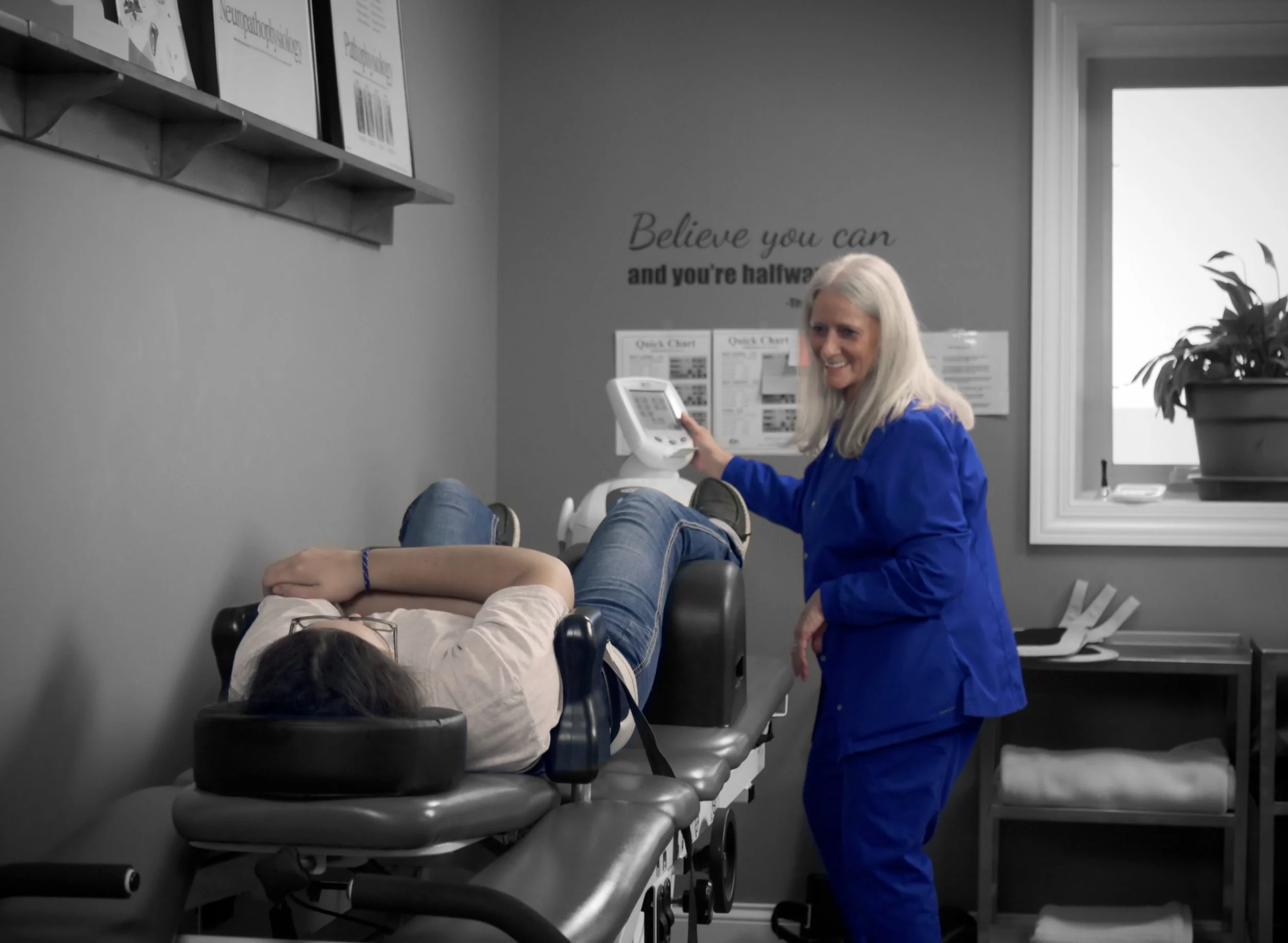 A woman lying on a medical examination table while a healthcare professional adjusts a medical device, the room has gray walls, a window with a potted plant, and inspirational wall decor.