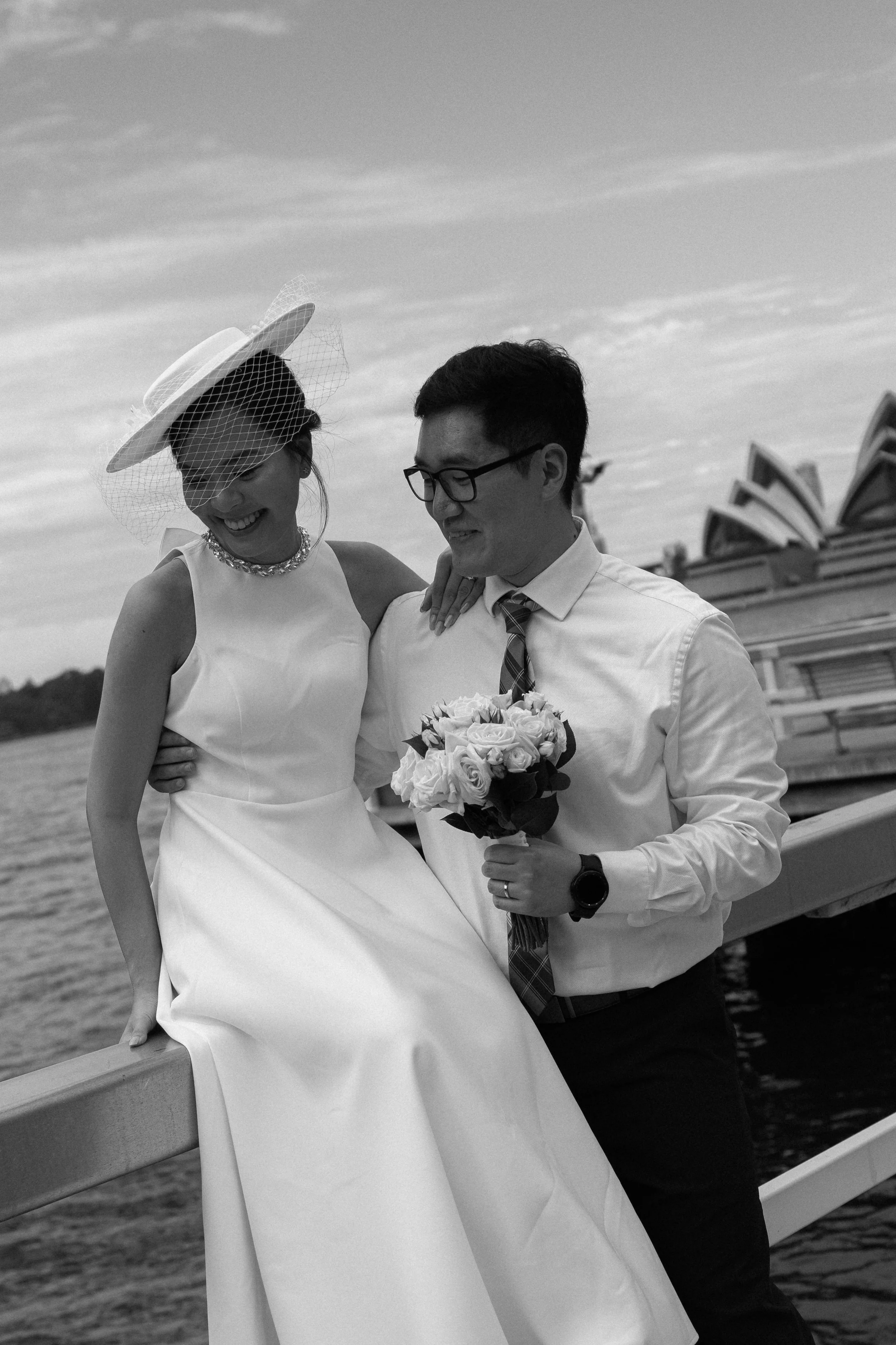 A black-and-white photo of a bride and groom by the water, smiling and embracing, with the Sydney Opera House in the background.