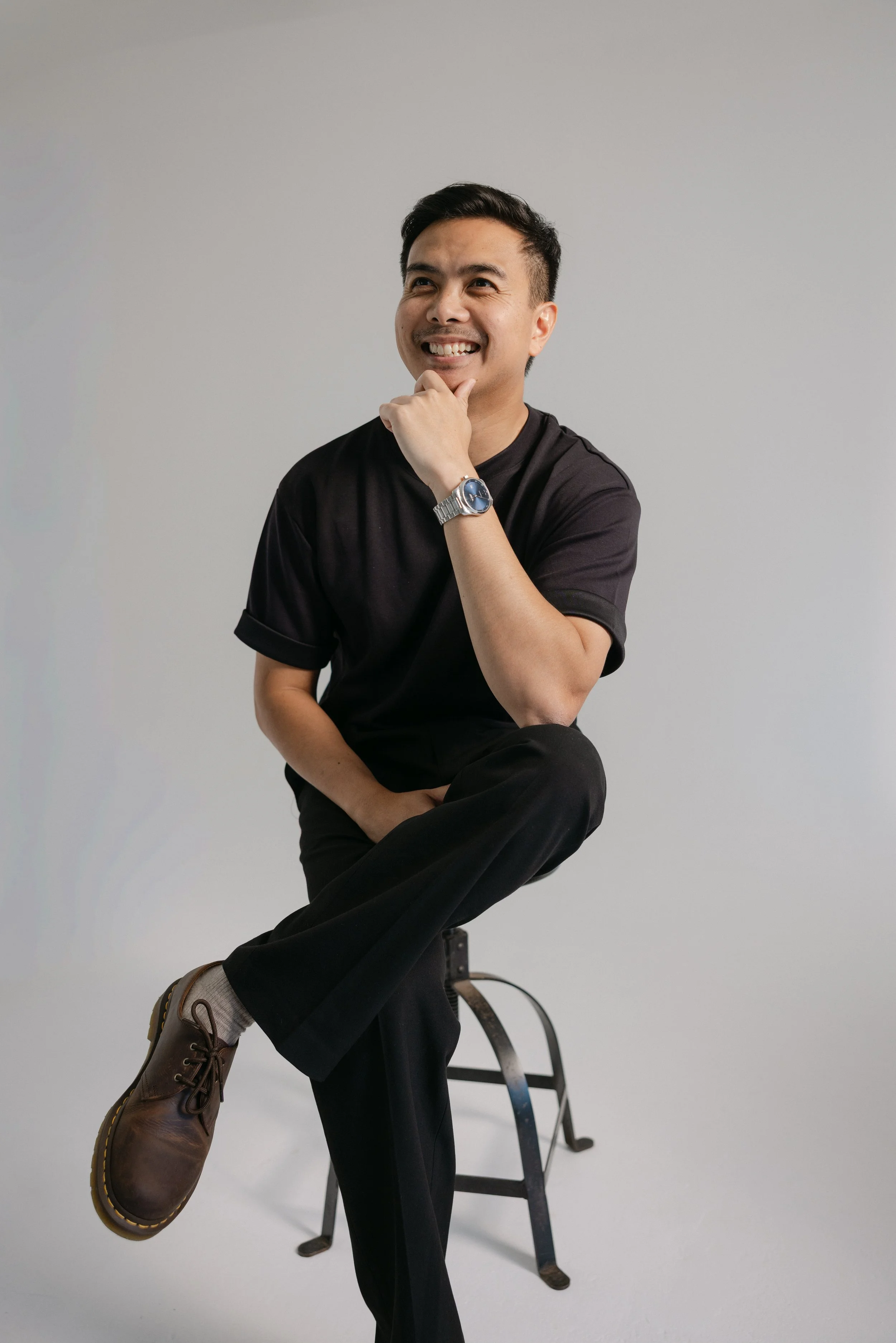 A man sitting on a stool, smiling, with his right hand on his chin, wearing a black t-shirt, black pants, brown shoes, and a wristwatch, against a plain light background.