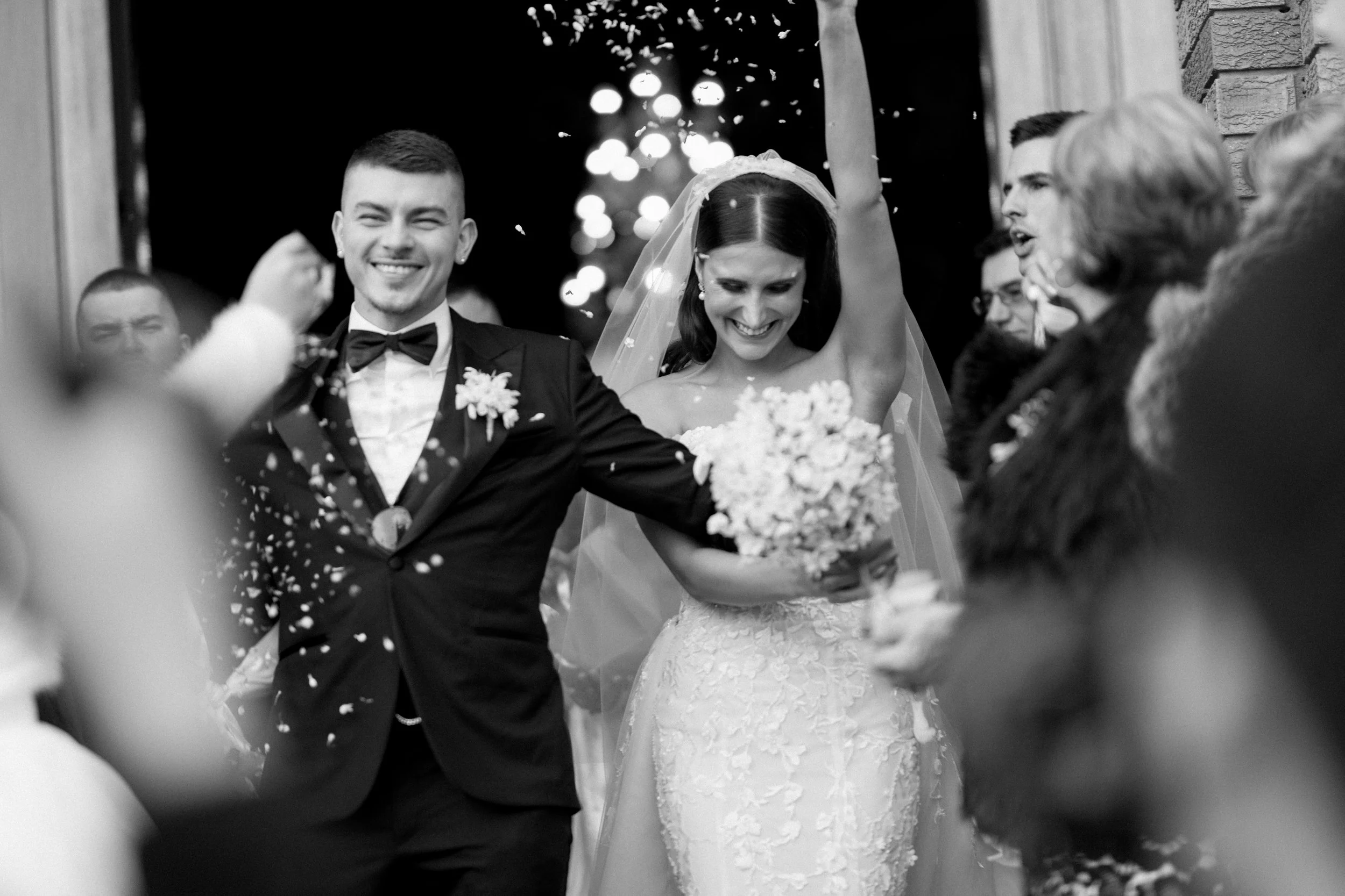 A bride and groom smiling as they leave a wedding ceremony, surrounded by guests with confetti falling.