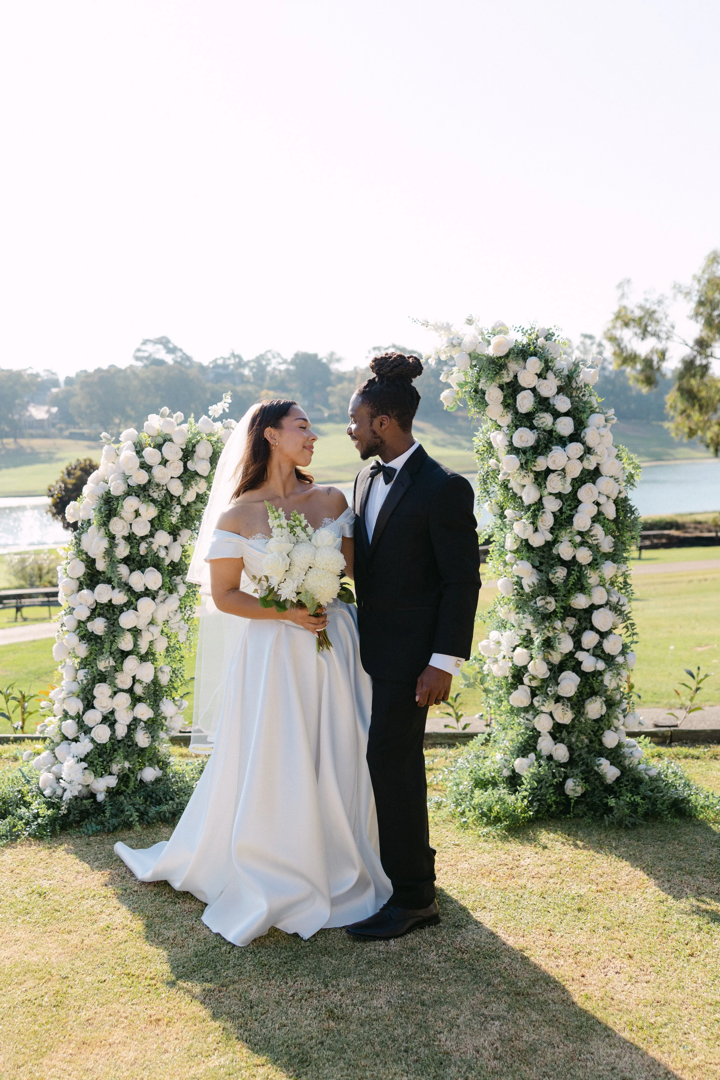 A newlywed couple on their wedding day standing close together, smiling at each other, framed by two large floral arrangements of white roses and greenery outdoors near a lake on a sunny day.