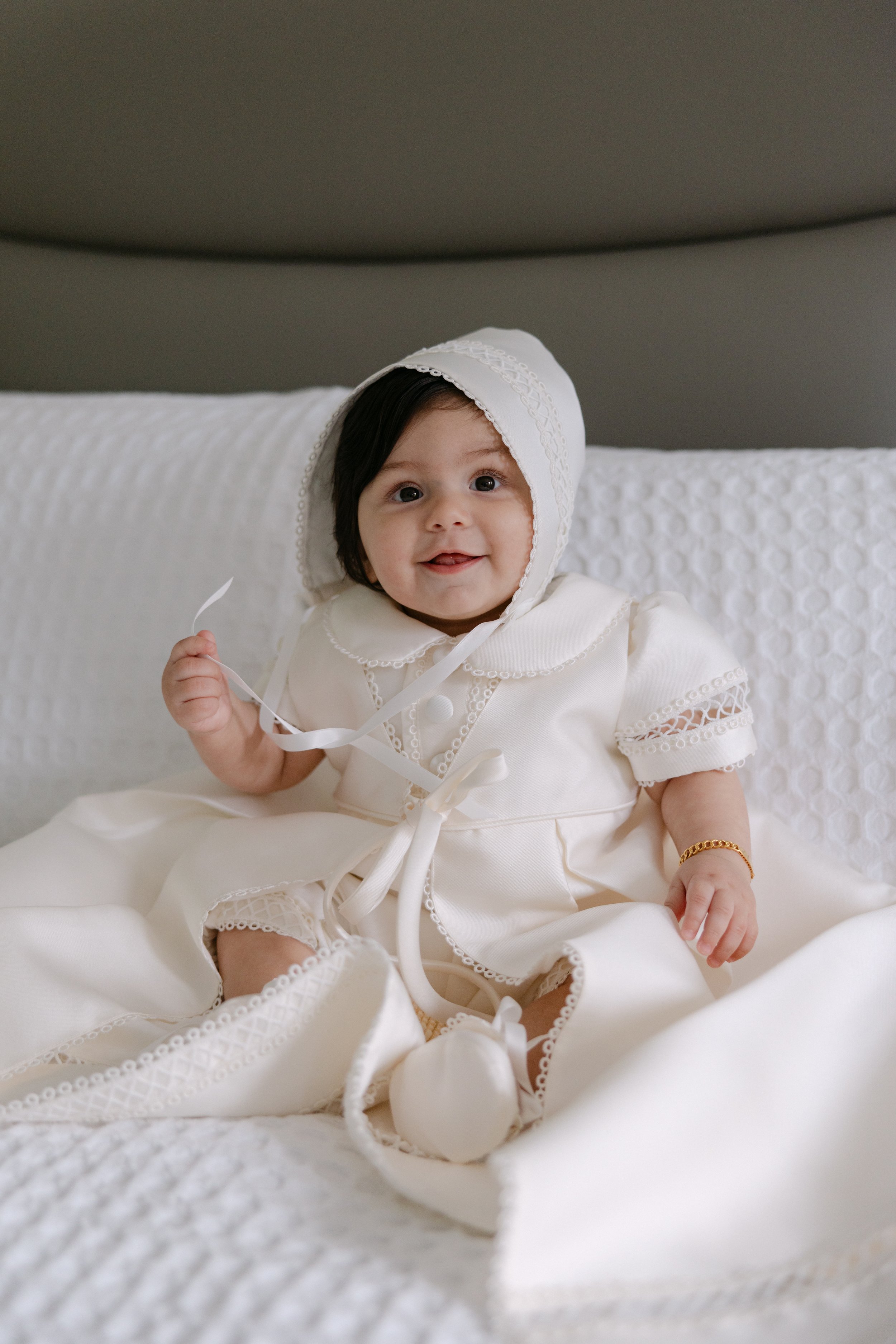 A smiling baby girl smiling at the camera, dressed in a white christening gown with lace details, sitting on a bed with white bedding.