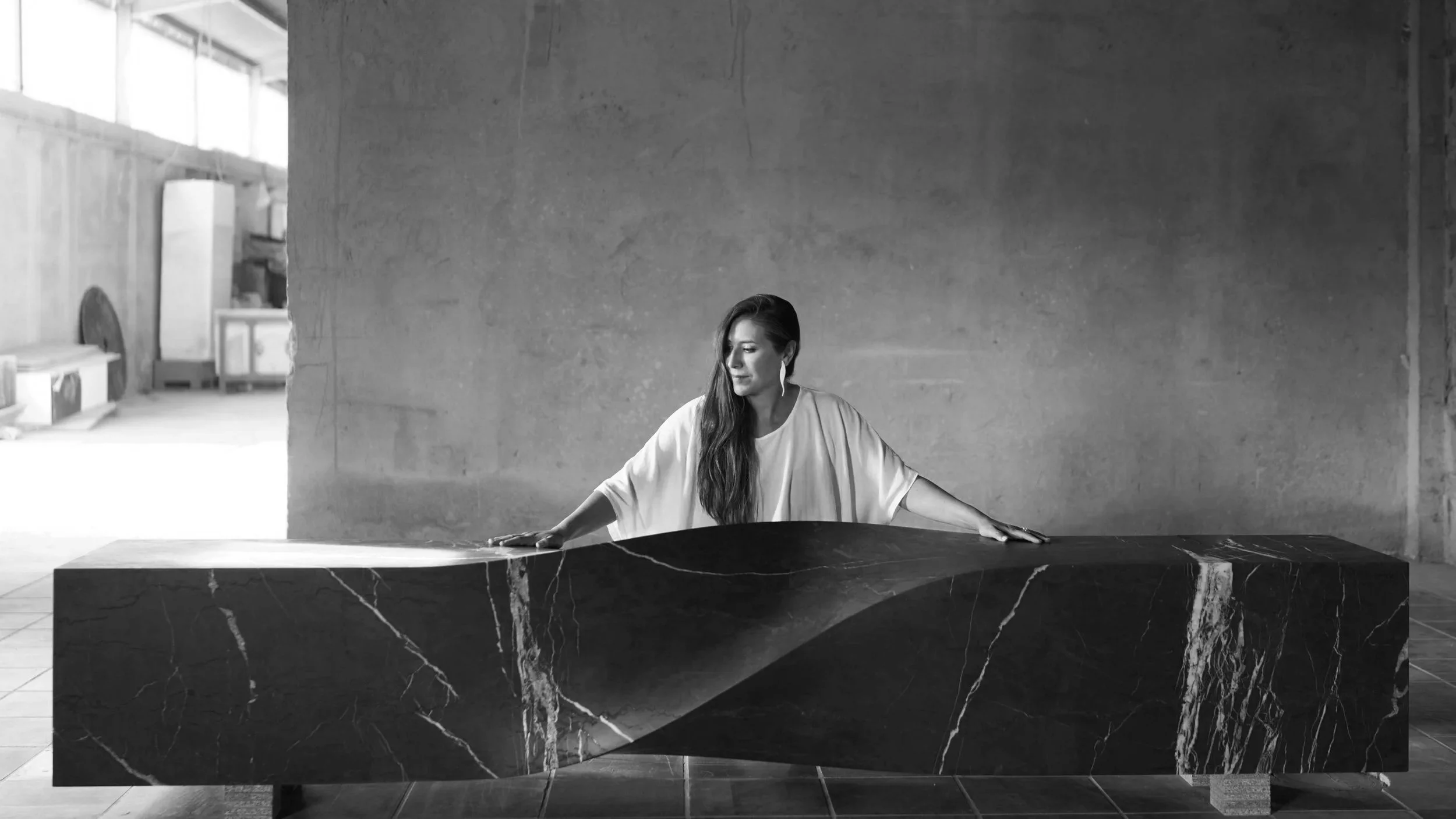 Woman in white resting her hands on a dark veined marble piece with a sculpted wave, in a black-and-white workshop portrait.