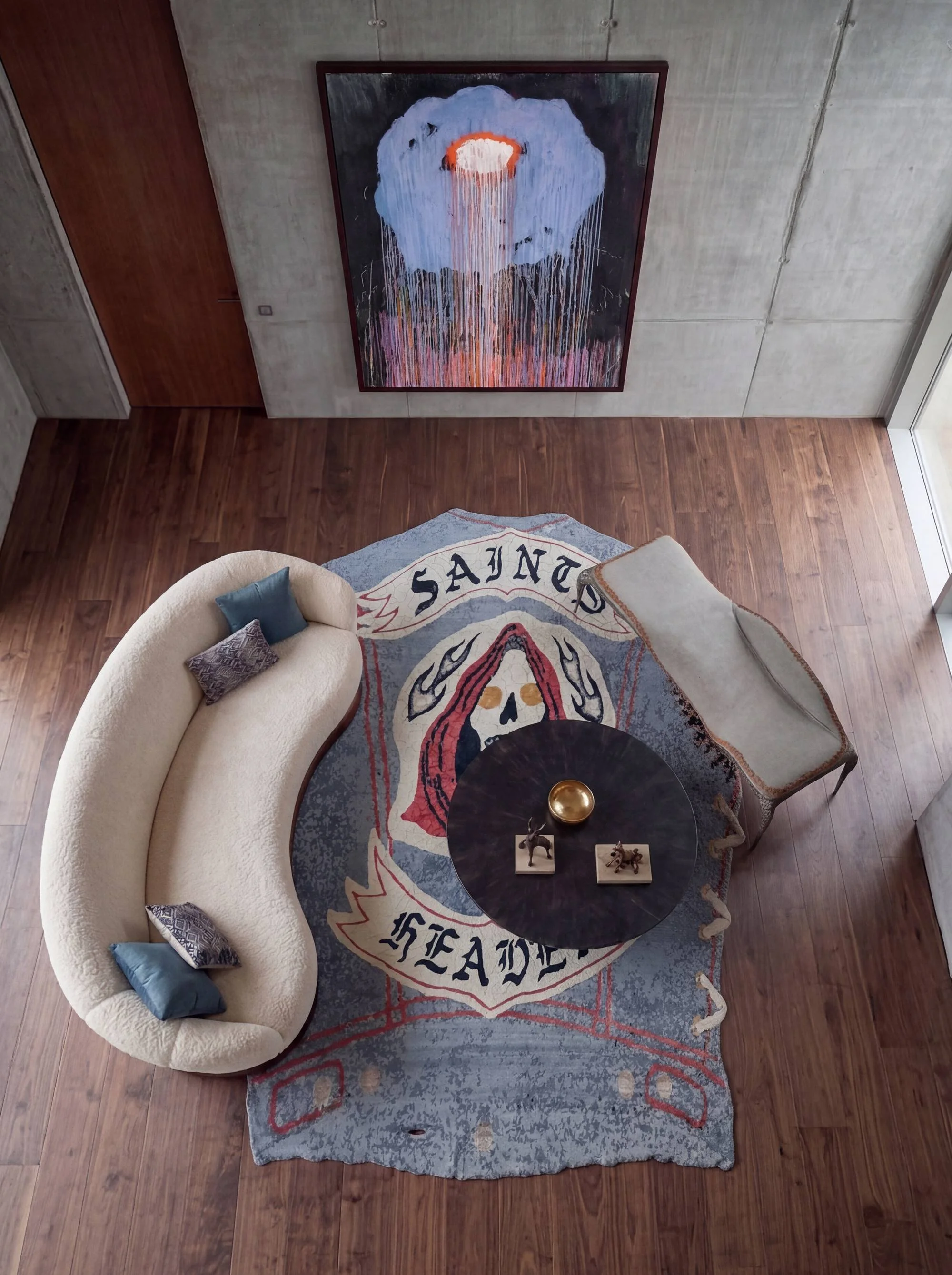Aerial view of a vest-shaped Dinodo Studio artist rug with a skull and gothic lettering, in a concrete living room with a curved sofa and black table.
