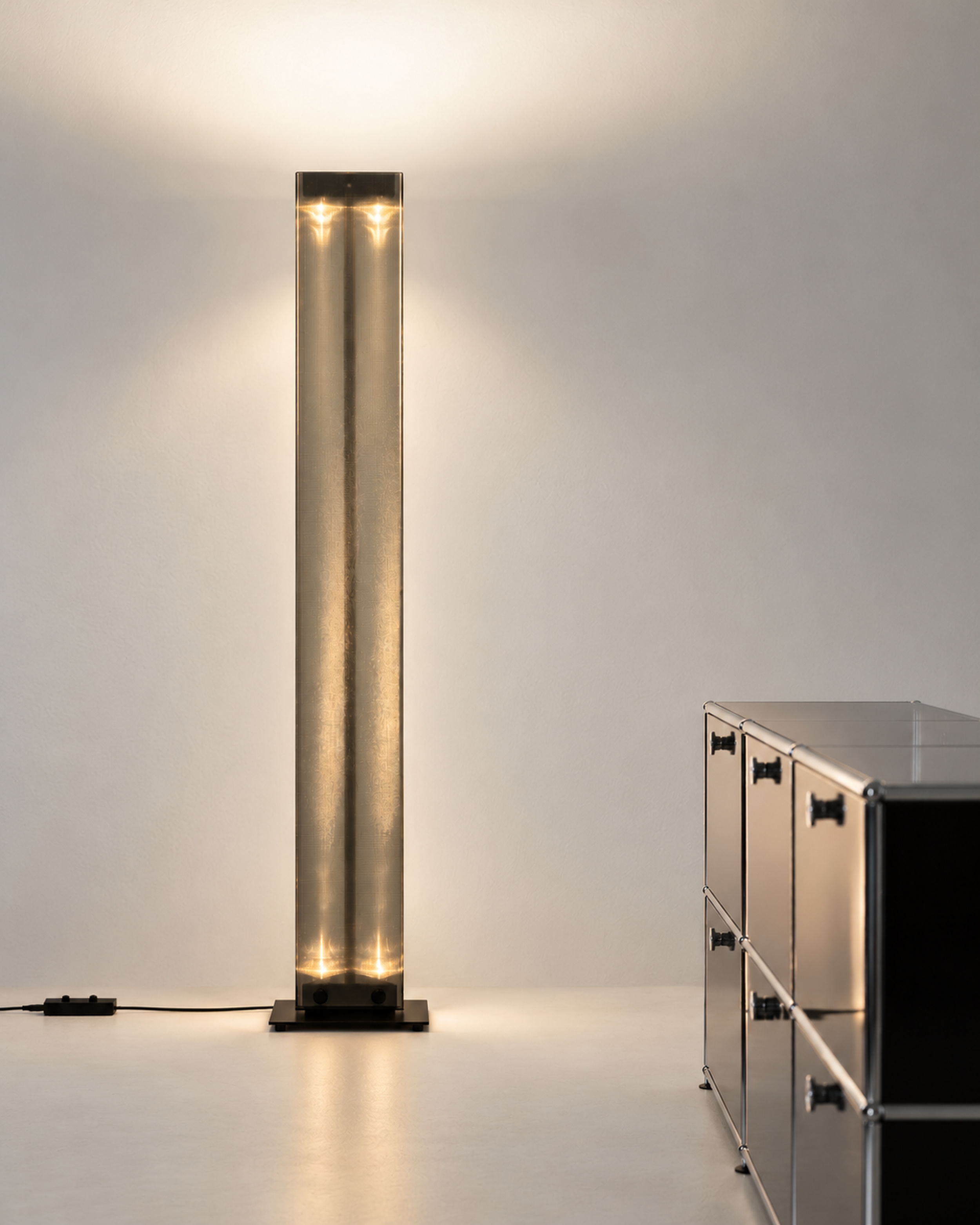 Belux Twilight floor lamp in black casting warm indirect light on a white wall beside a steel sideboard