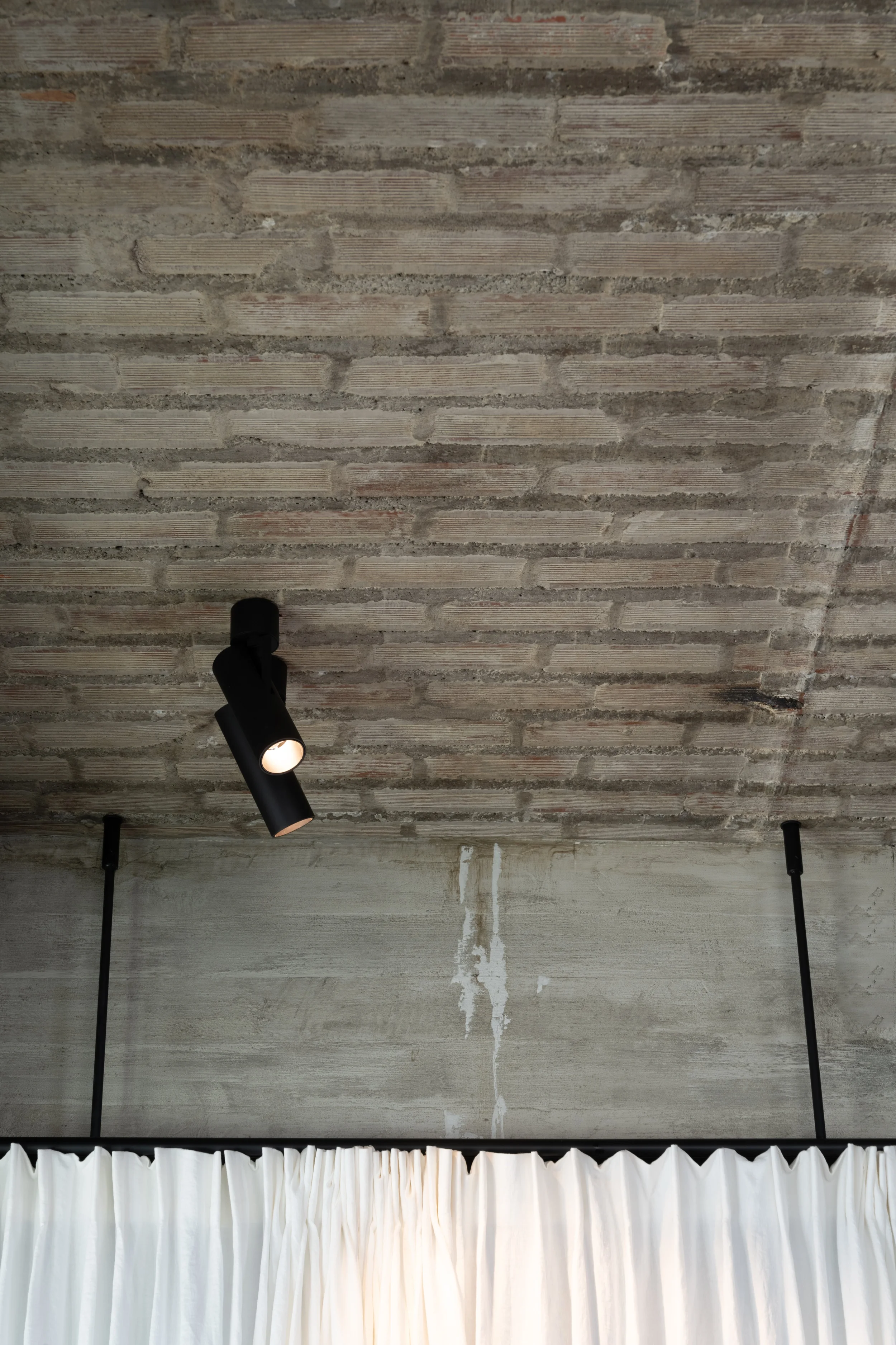 Black Kreon Holon 60 Directional Surface spotlight on a brick-patterned concrete ceiling above a linen curtain