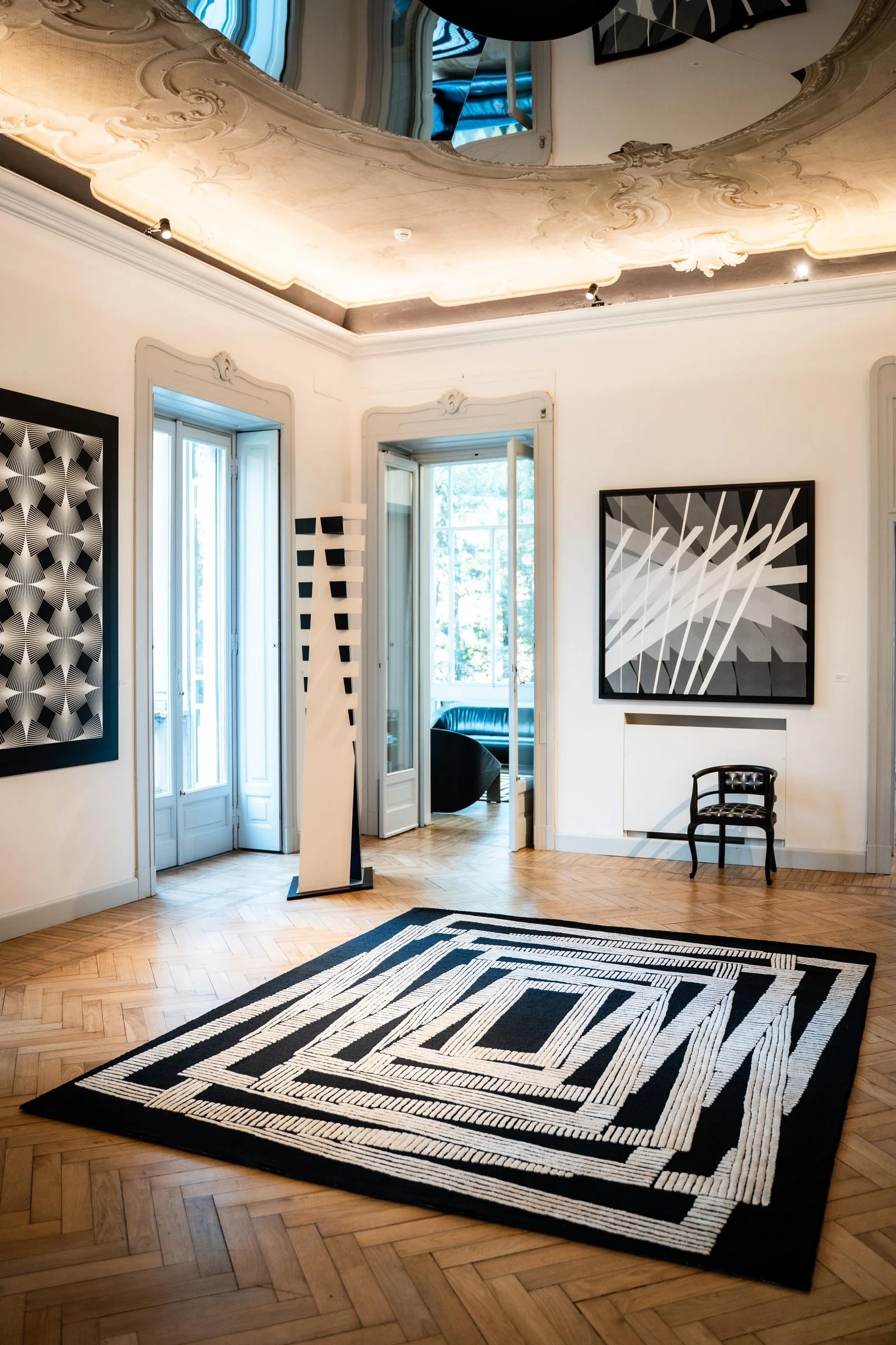 Black and white Dinodo Studio artist rug with concentric rectangles, exhibited on a herringbone floor in a gallery with ornate ceilings and op art works.