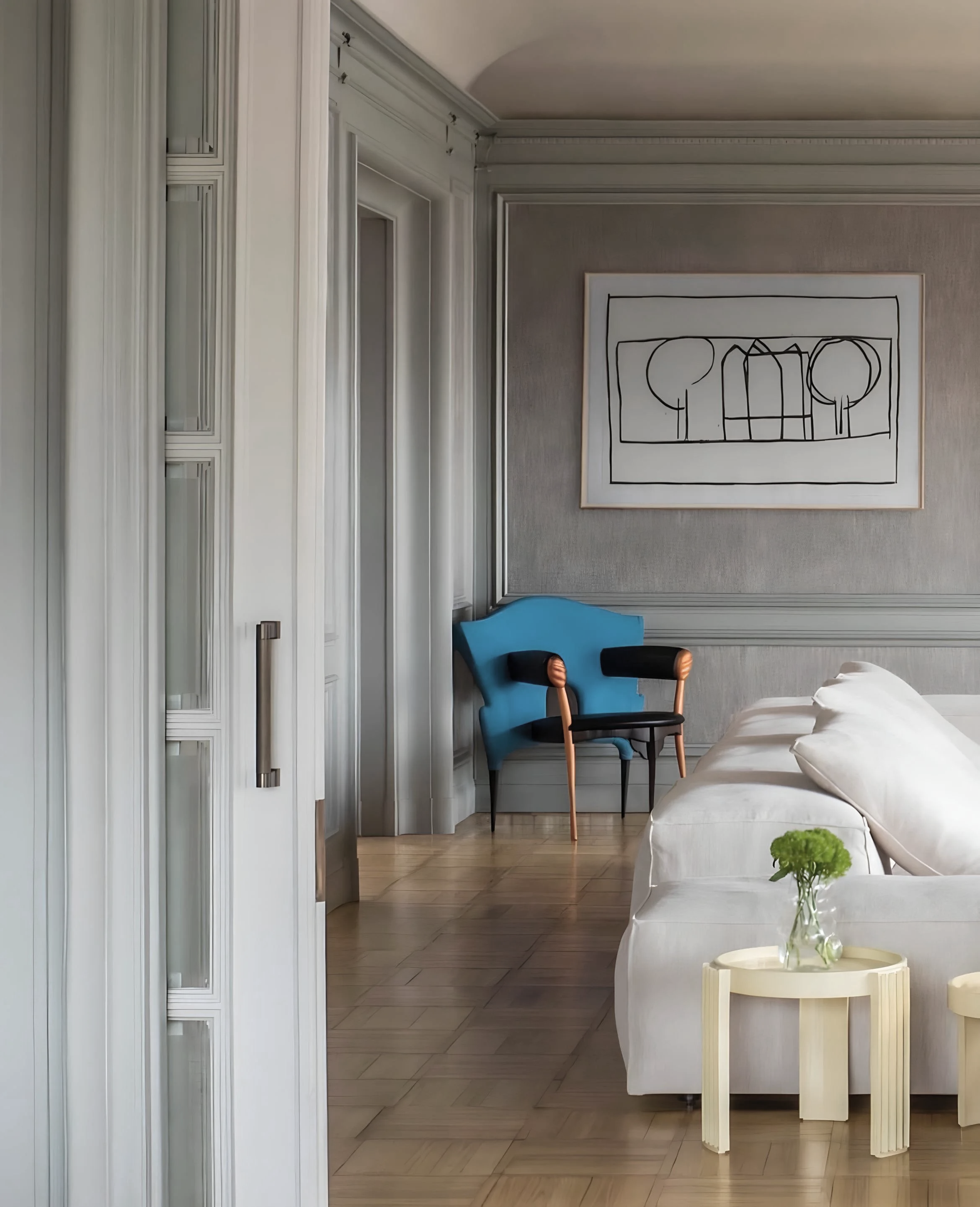 Grey paneled room with a blue sculptural armchair, a line drawing of trees and houses, a white sofa and a small side table.