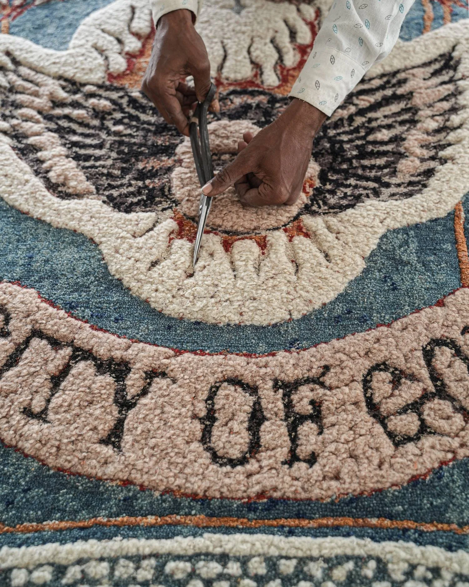 Artisan hand-carving the pile of a hand-knotted Dinodo Studio rug with scissors, revealing wings, flames and lettering in cream, blue and red.