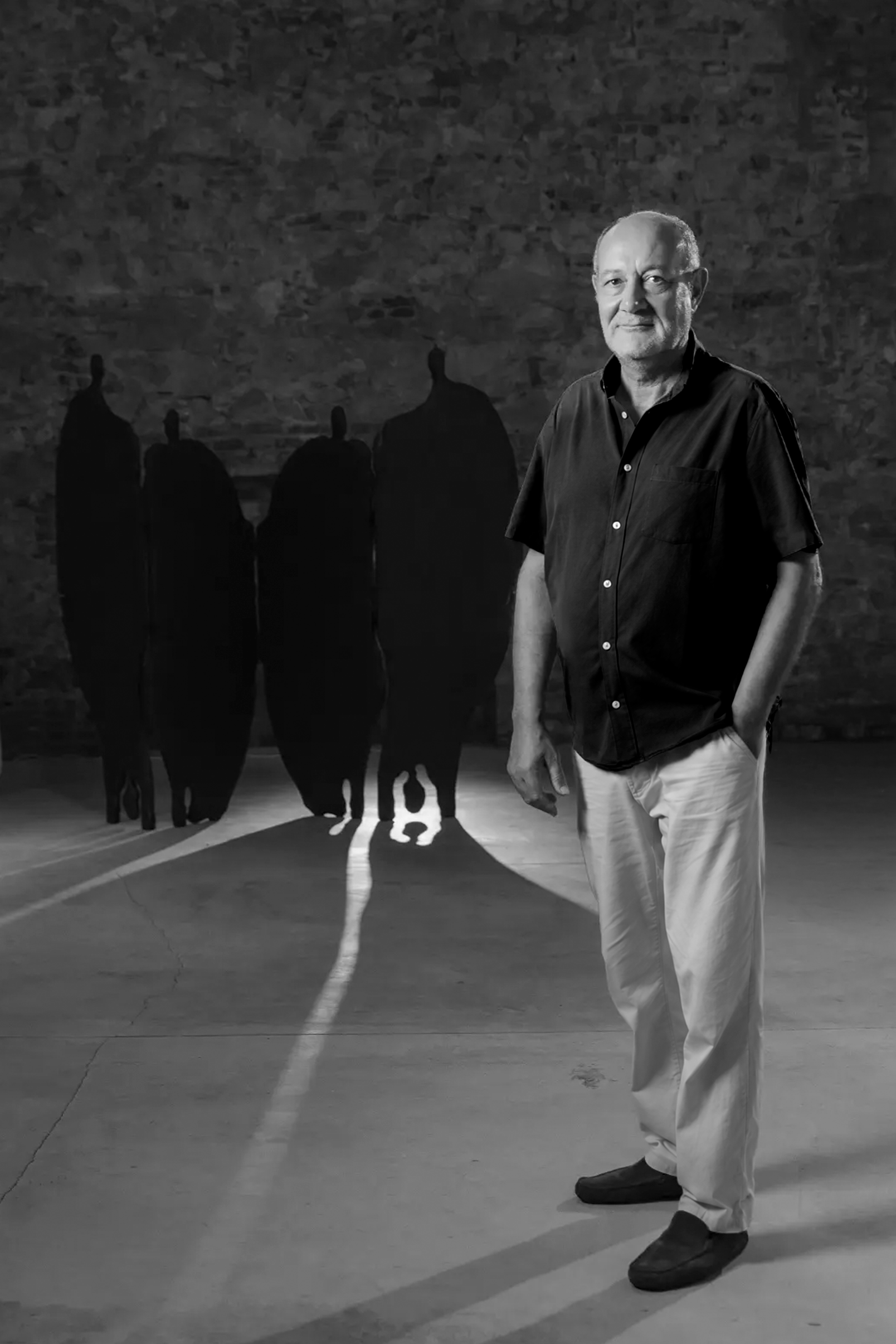 Robert Pous standing beside four tall dark figure sculptures, light spilling between them, in a black-and-white portrait.