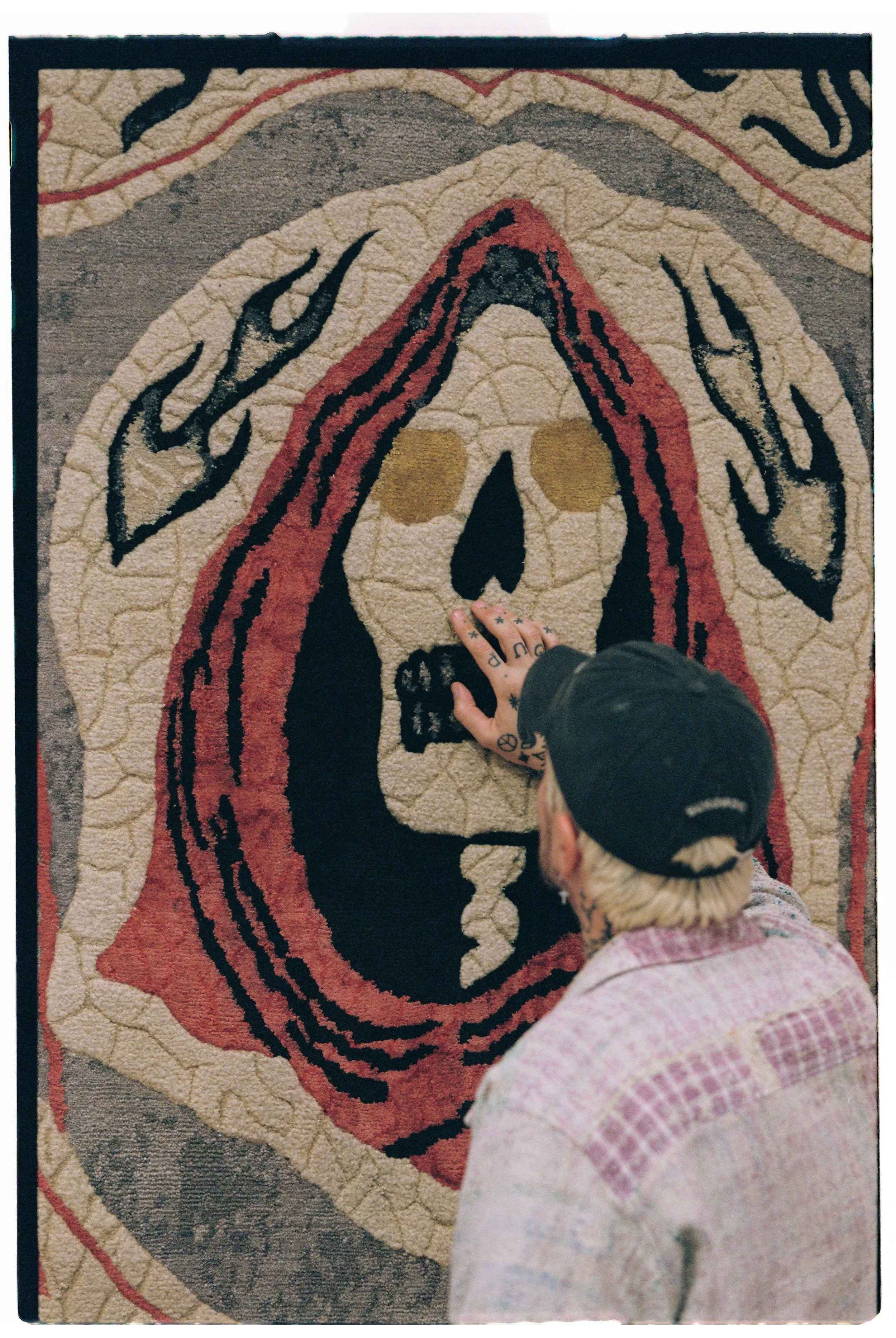 Artist Julien Jaca touching the hooded skull of his Saints hand-knotted rug for Dinodo Studio, hung on the wall as a tapestry, available on BLANCONEGRO BARCELONA
