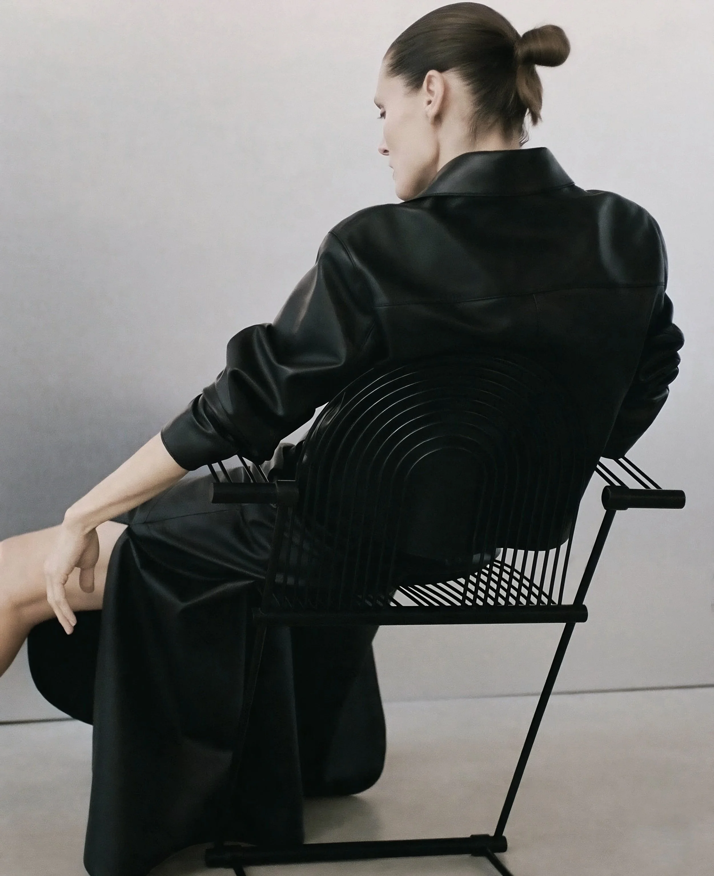 Woman in black leather seated on a black metal chair with a concentric arched rod backrest, against a soft grey backdrop.