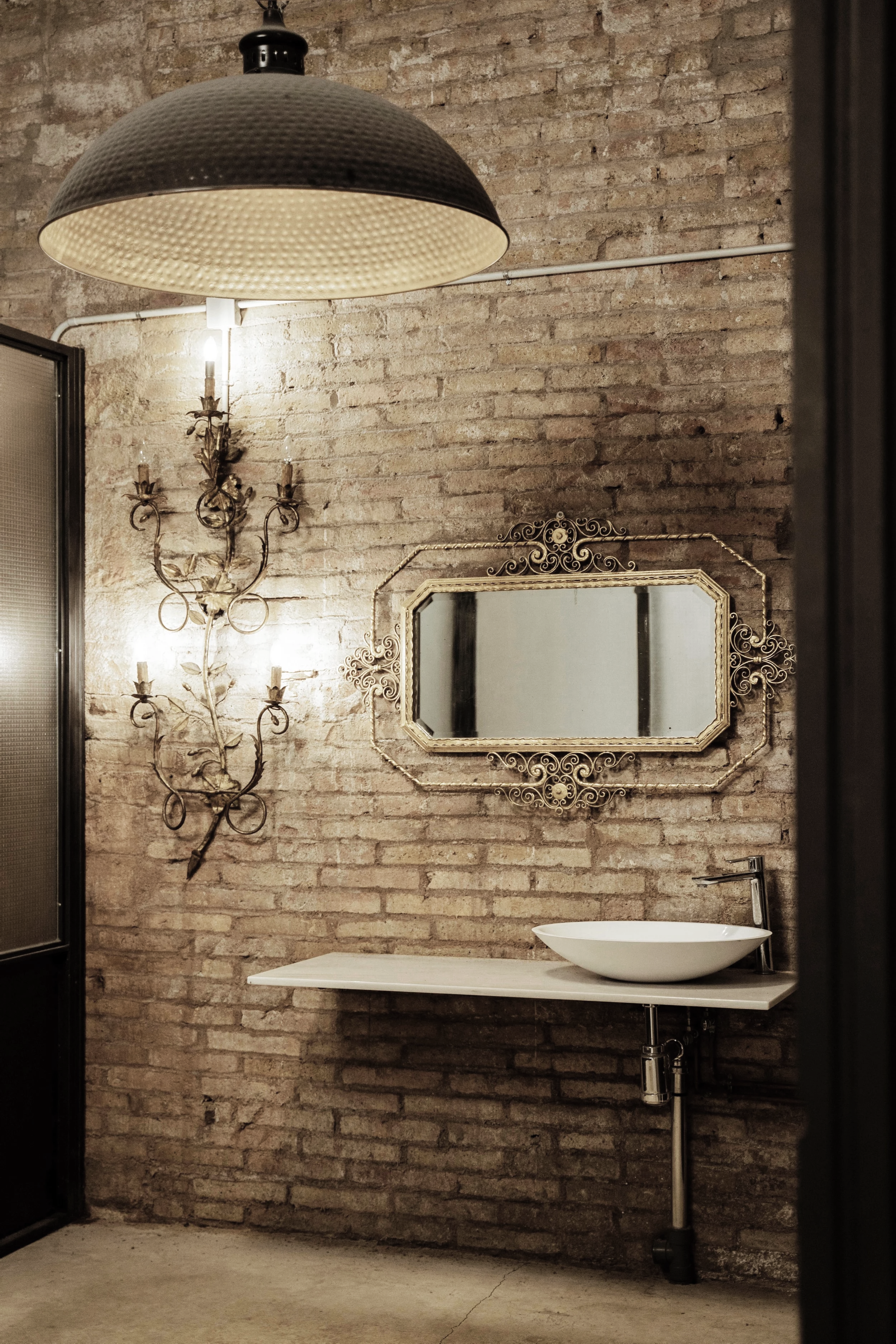 BLANCONEGRO bathroom: exposed brick, ornate gilt candelabra sconce, filigree mirror, white vessel sink and hammered pendant.