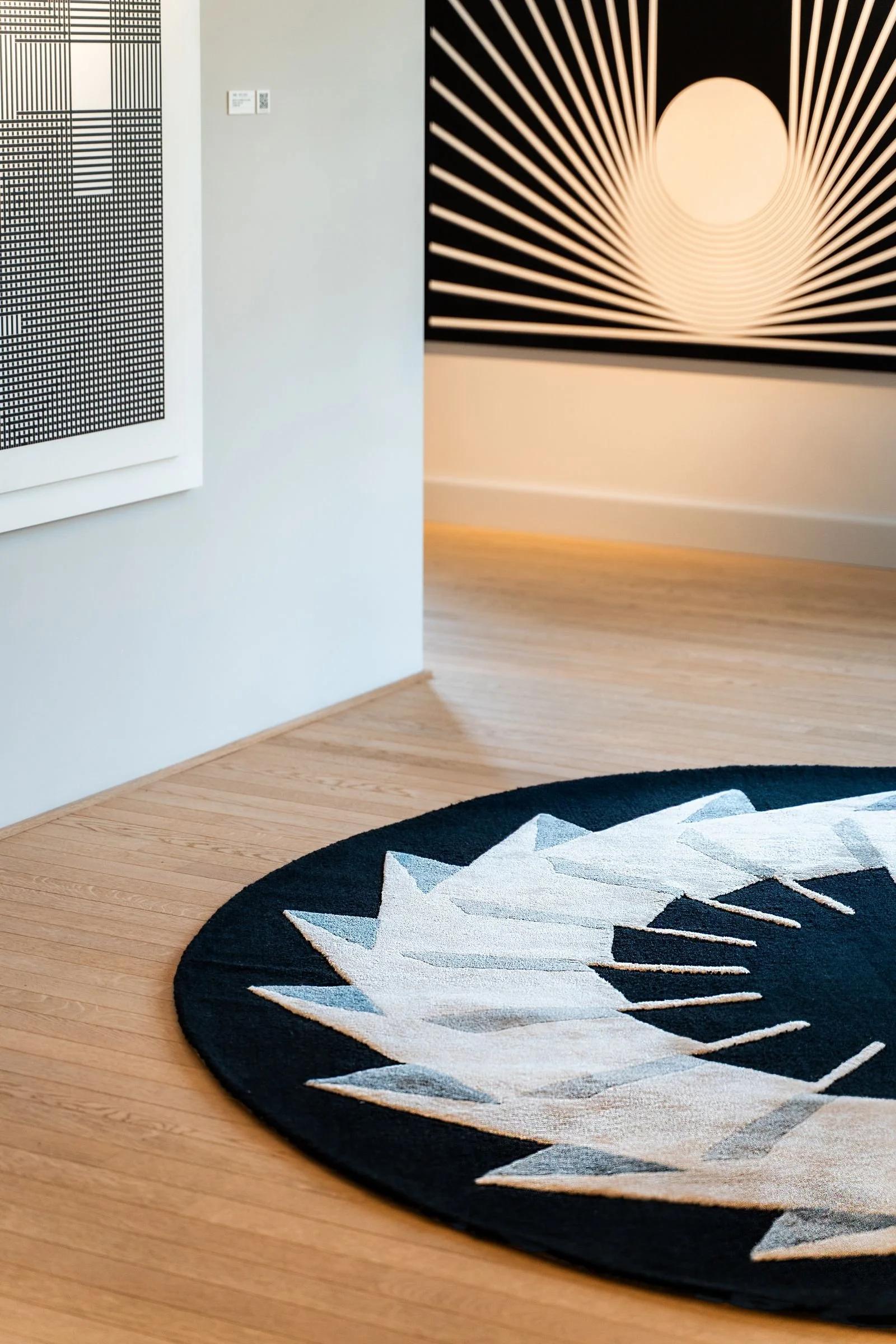 Round Dinodo Studio artist rug in black, cream and grey with geometric shapes, exhibited in an art gallery with a wooden floor.