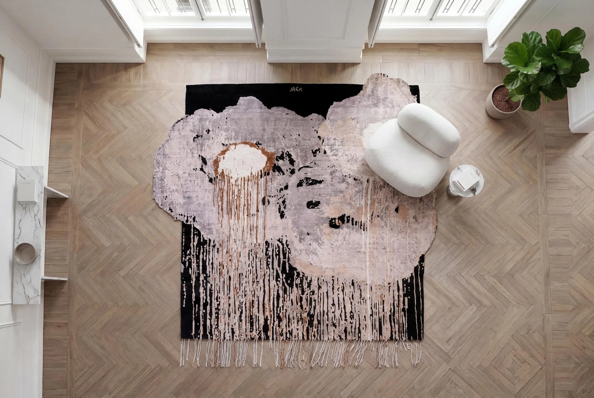 Aerial view of a Dinodo Studio artist rug with an organic shape, drips and fringes in black, pink and cream, on a herringbone floor beside a white armchair.