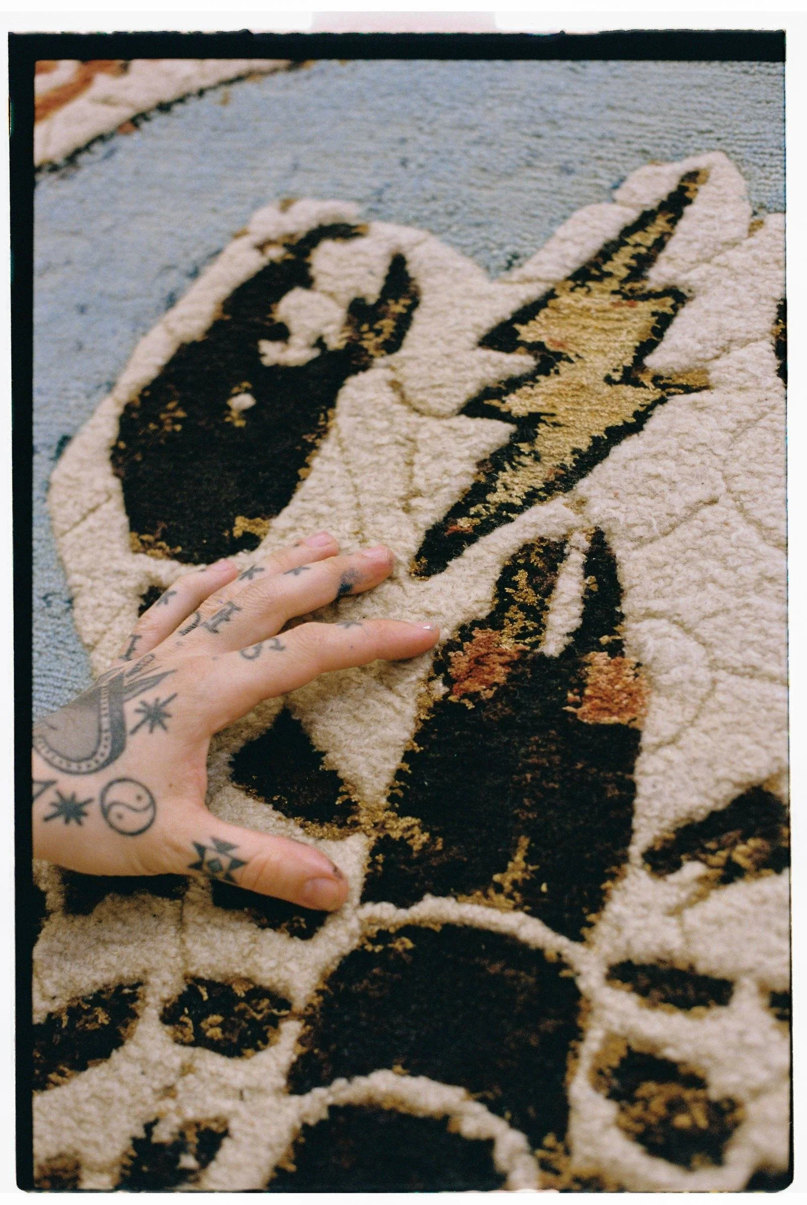 Tattooed hand touching a hand-knotted Dinodo Studio rug: close-up of the pile texture in cream, black, gold and blue with a lightning bolt.