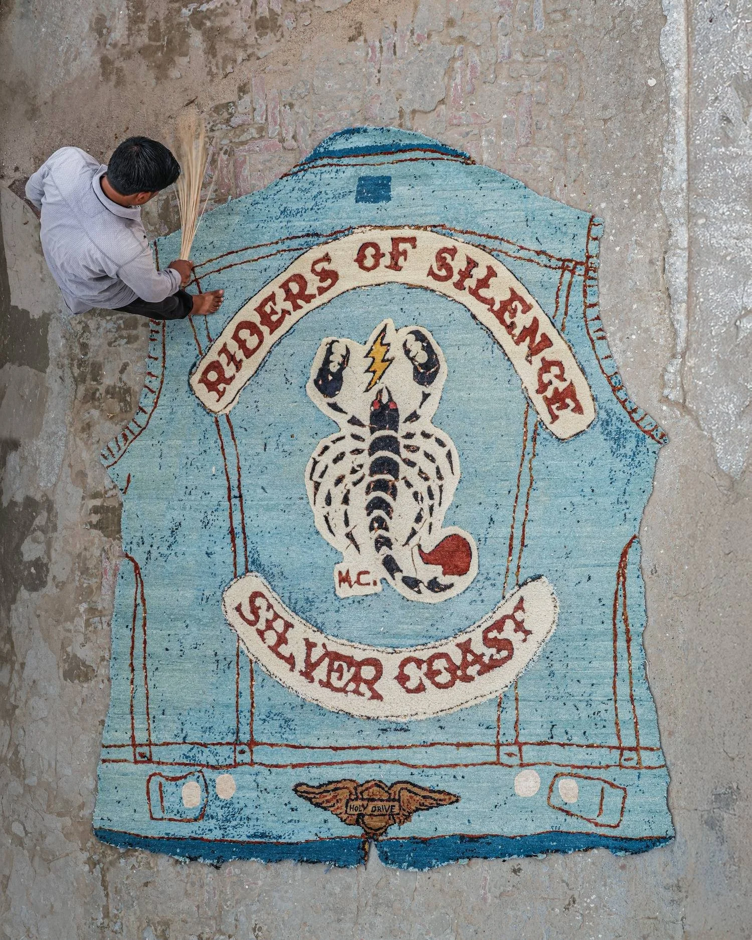 Aerial view of an artisan finishing a Dinodo Studio artist rug shaped like a denim vest, brushing the pile by hand with a natural fiber broom.