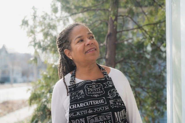 Woman with braided hair wearing a black apron with white text, standing outdoors near trees.