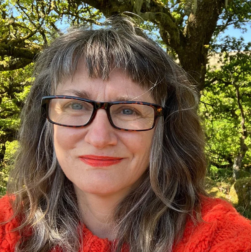 Close-up photo of a woman with gray and brown curly hair, wearing glasses and red lipstick, smiling outdoors with green trees and blue sky in the background.