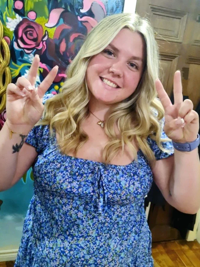 Smiling woman with long blonde hair, wearing a blue floral dress, holding up peace signs in front of a colorful mural.