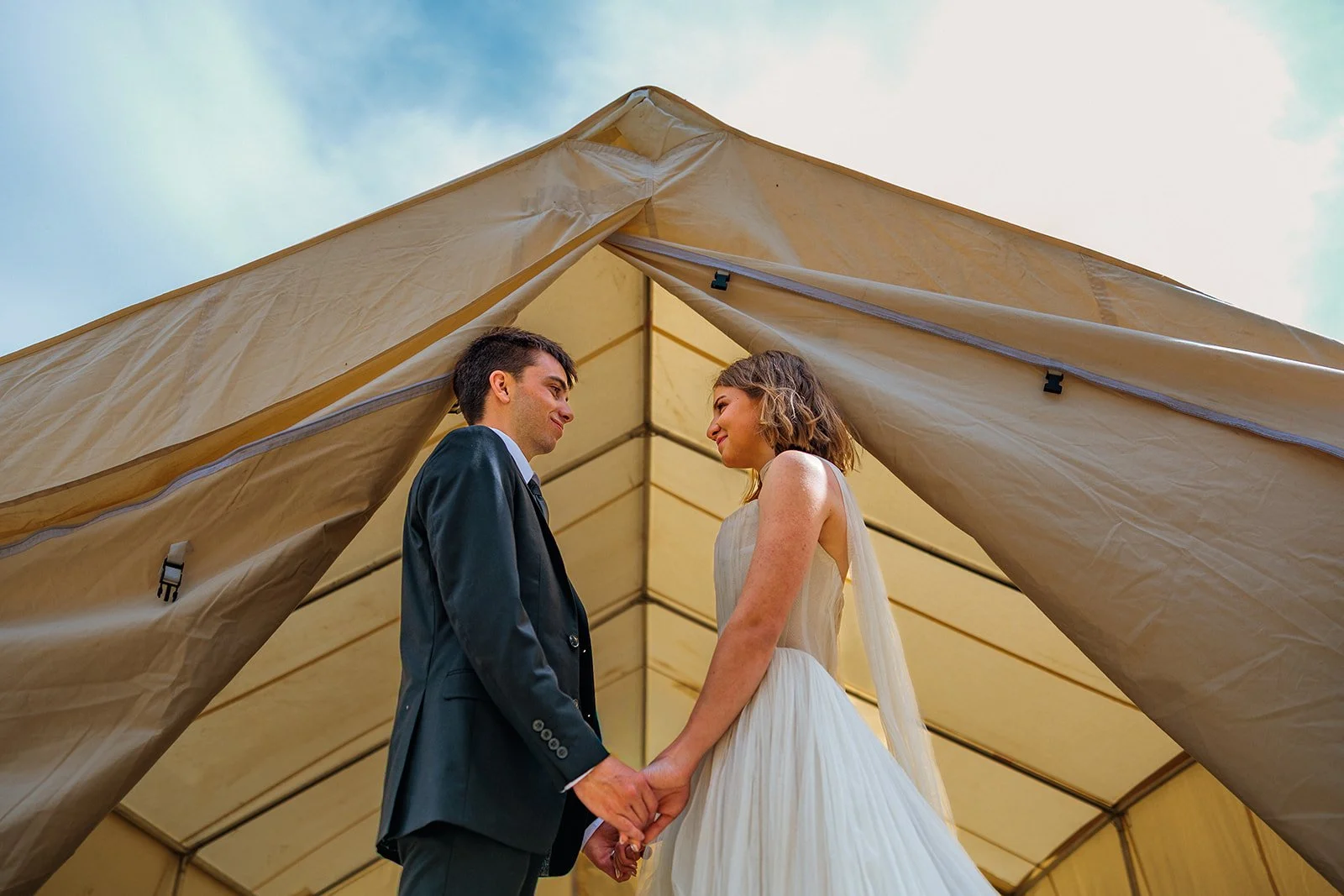 A bride and groom holding hands and gazing at each other inside a large tent during their wedding ceremony.