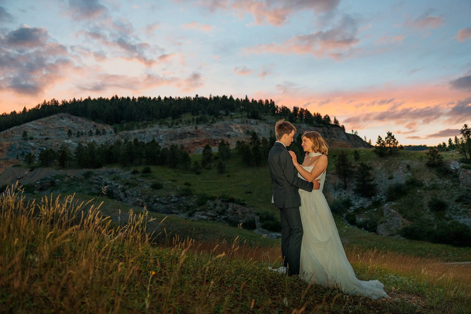 A newlywed couple stands outdoors during sunset, embracing each other and smiling, with a backdrop of rolling hills and a colorful sky.