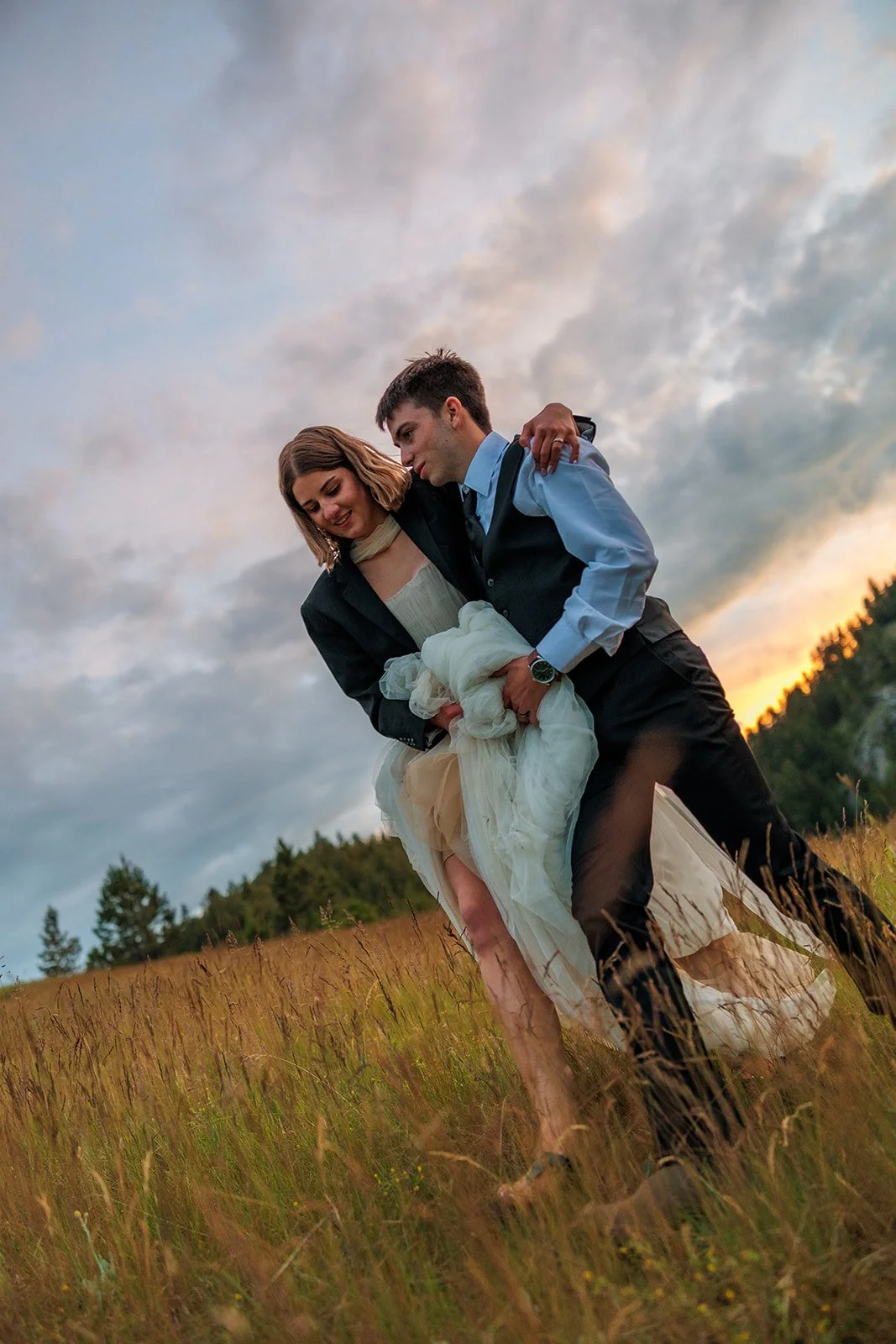 A couple in wedding attire standing in a field during sunset, with the groom holding the bride and both smiling.