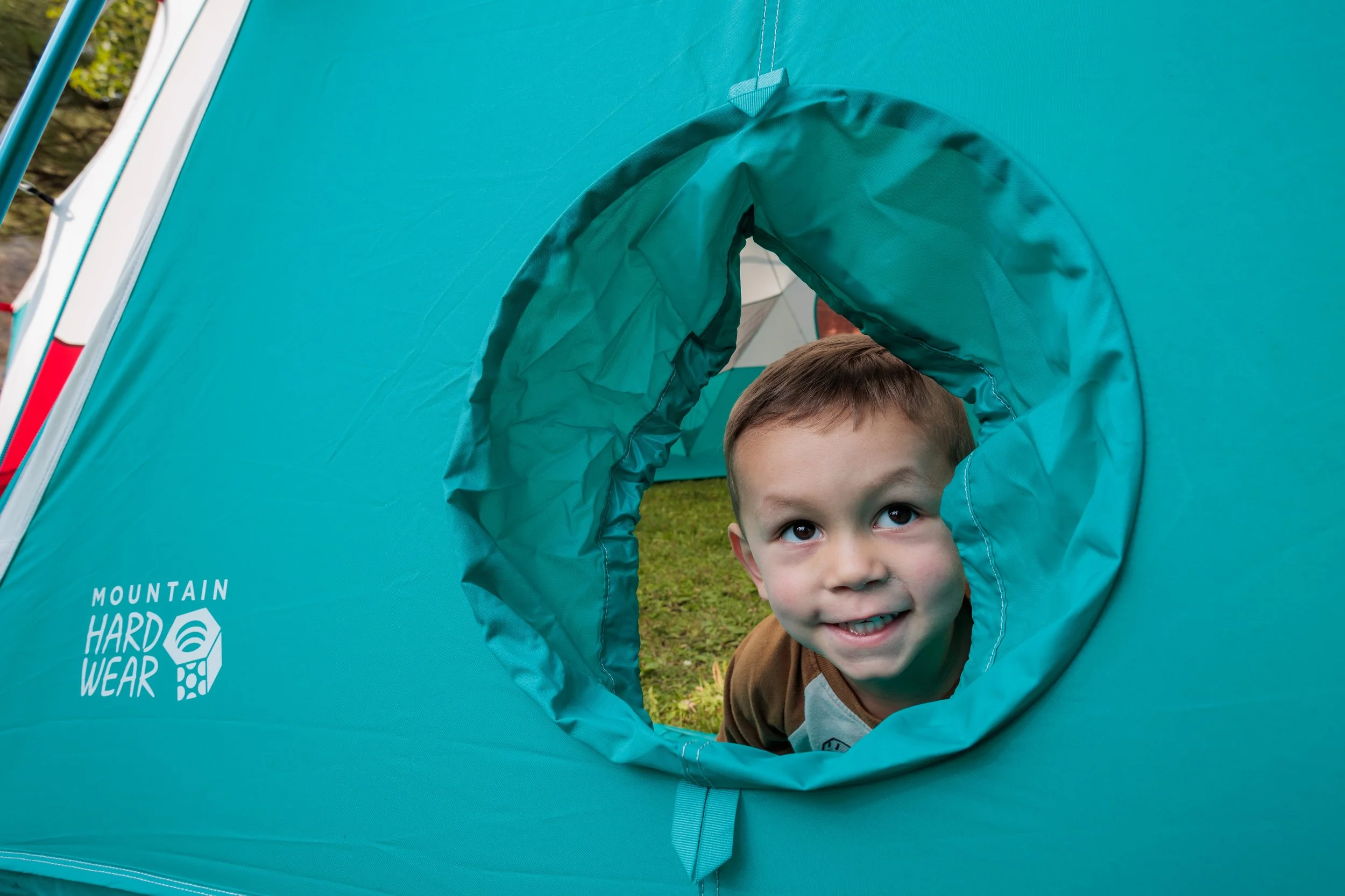 A young boy looking through a circular window in a teal tent on a grassy outdoor area.