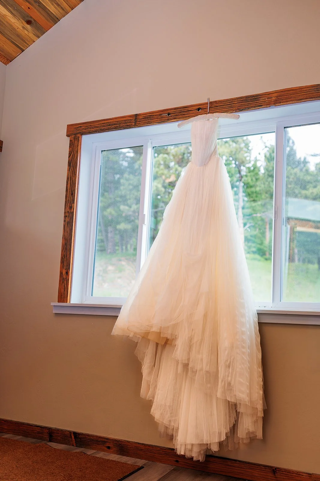 A wedding dress hanging from a hook in front of a window indoors.