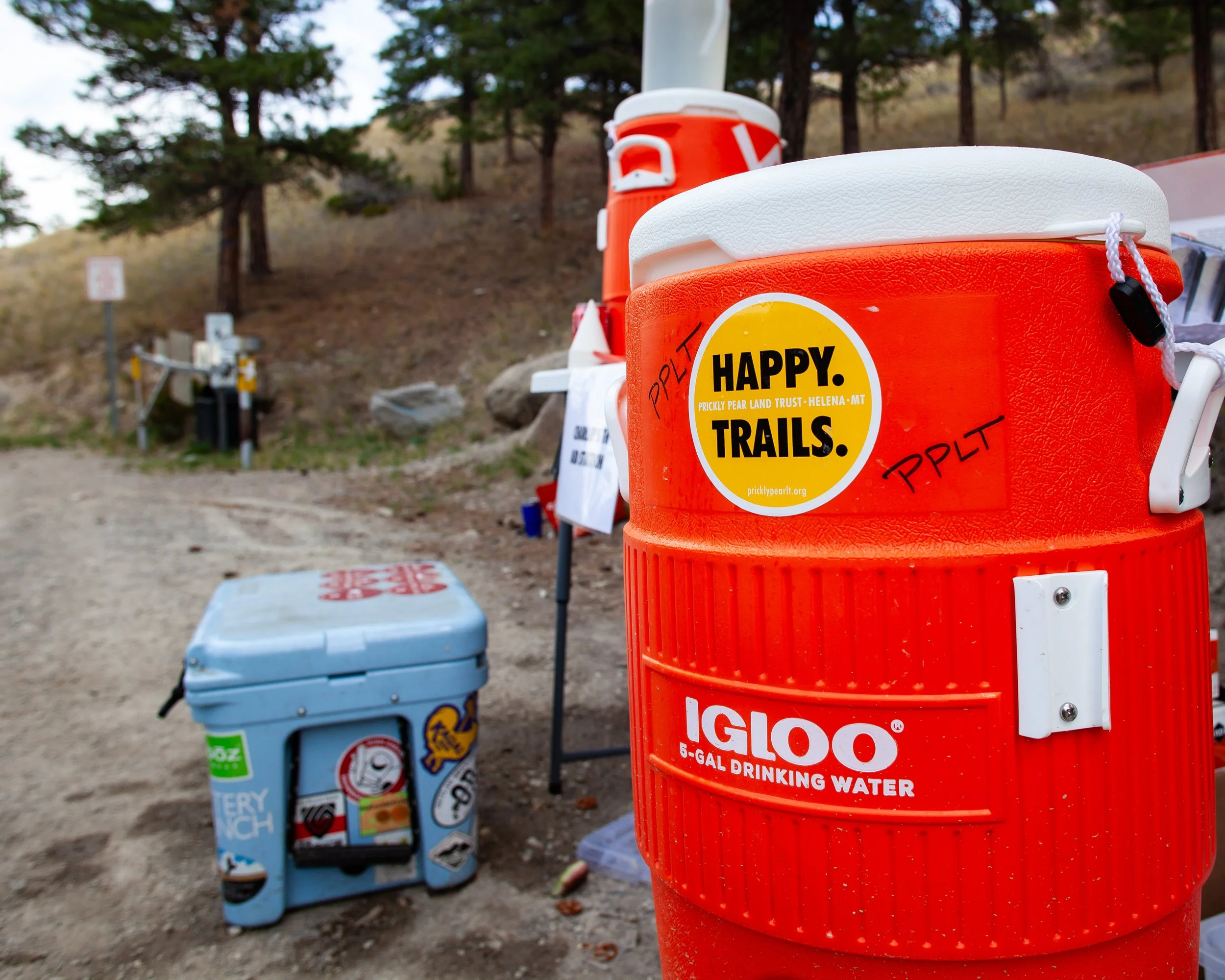 Close-up of orange Igloo water cooler with Happy Trails sticker, wooden sign with writing in the background, and a small cooler on dirt ground in a wooded outdoor trailhead area.