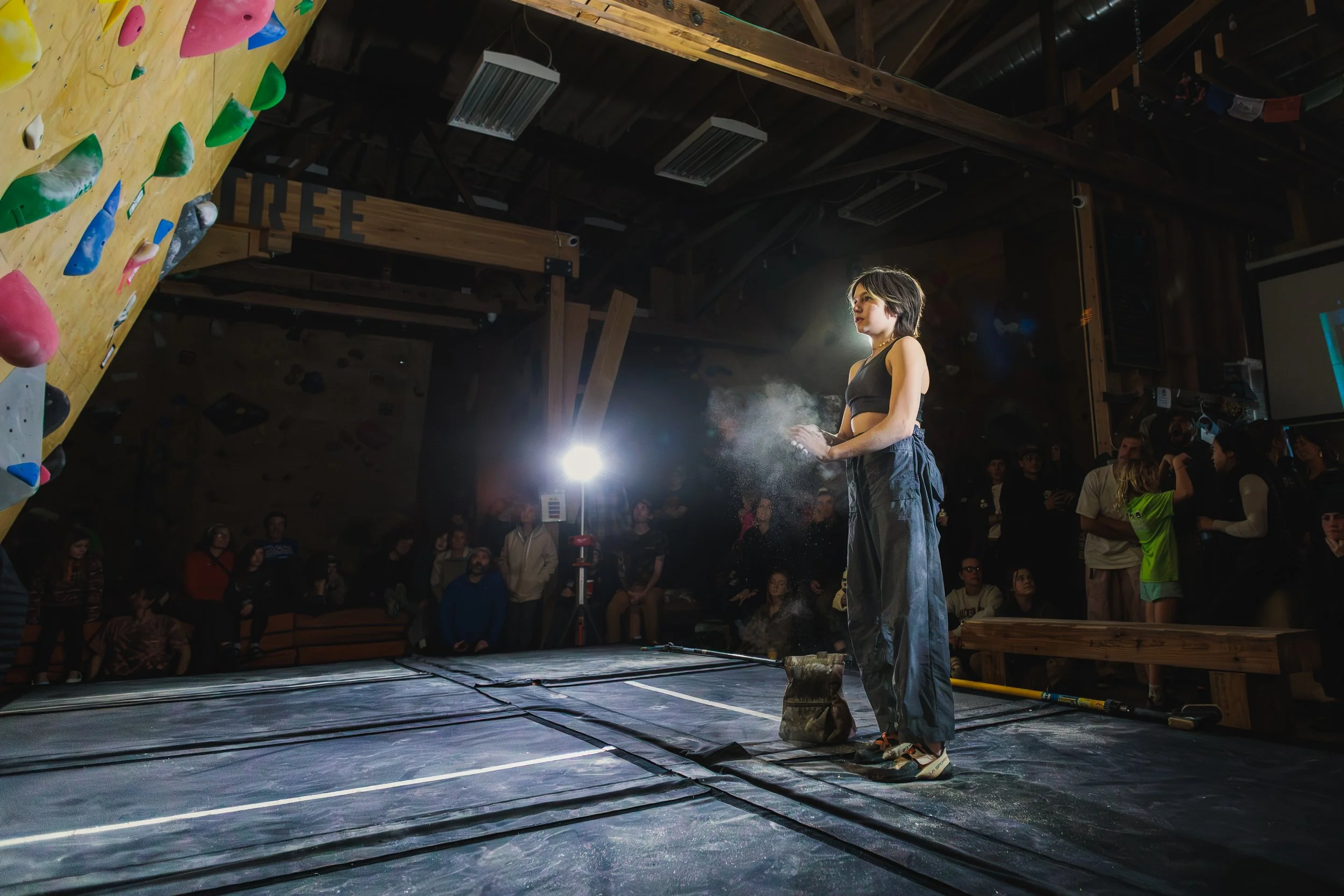 A woman preparing chalk for her climbing route on an indoor rock climbing wall with holds, while a crowd watches in a spacious indoor climbing gym.