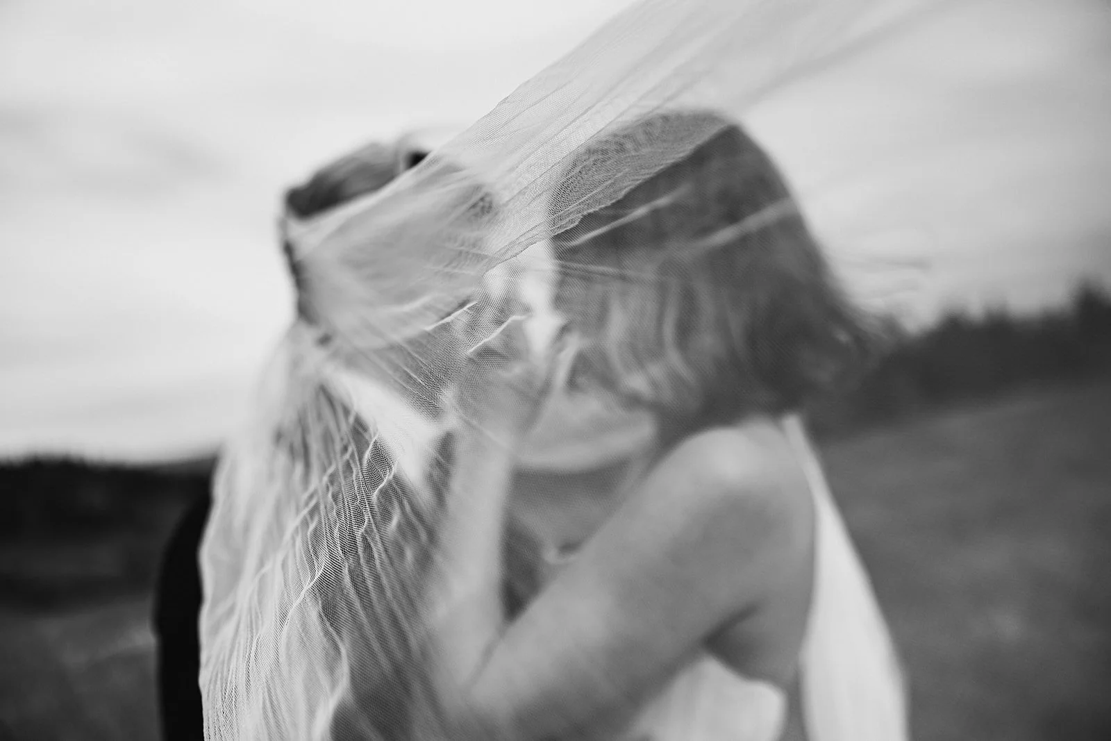 A person holding a veil over their face outdoors, background slightly blurred, in black and white.