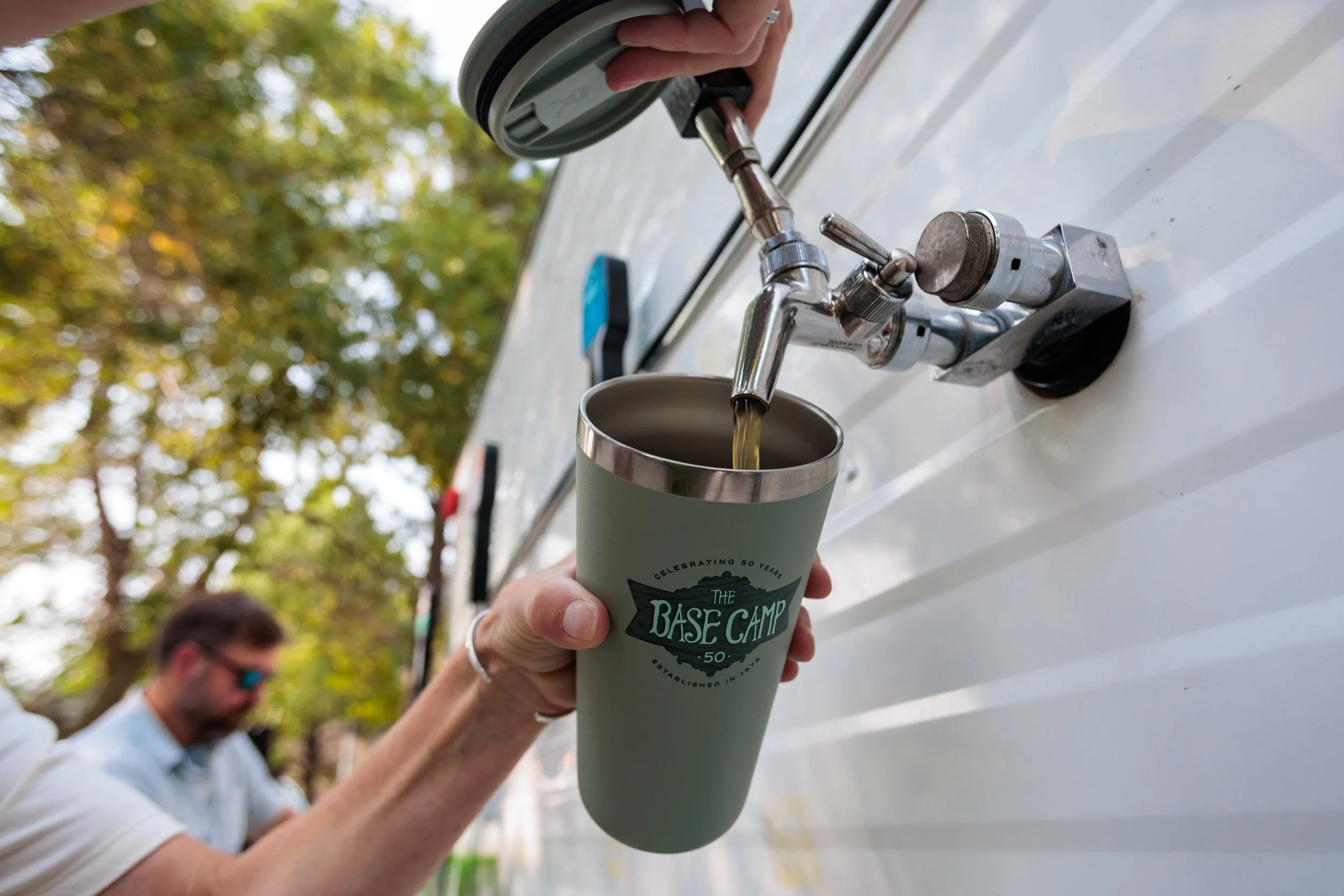 Person filling a green reusable cup with water from an outdoor spigot, with trees and another person in the background.