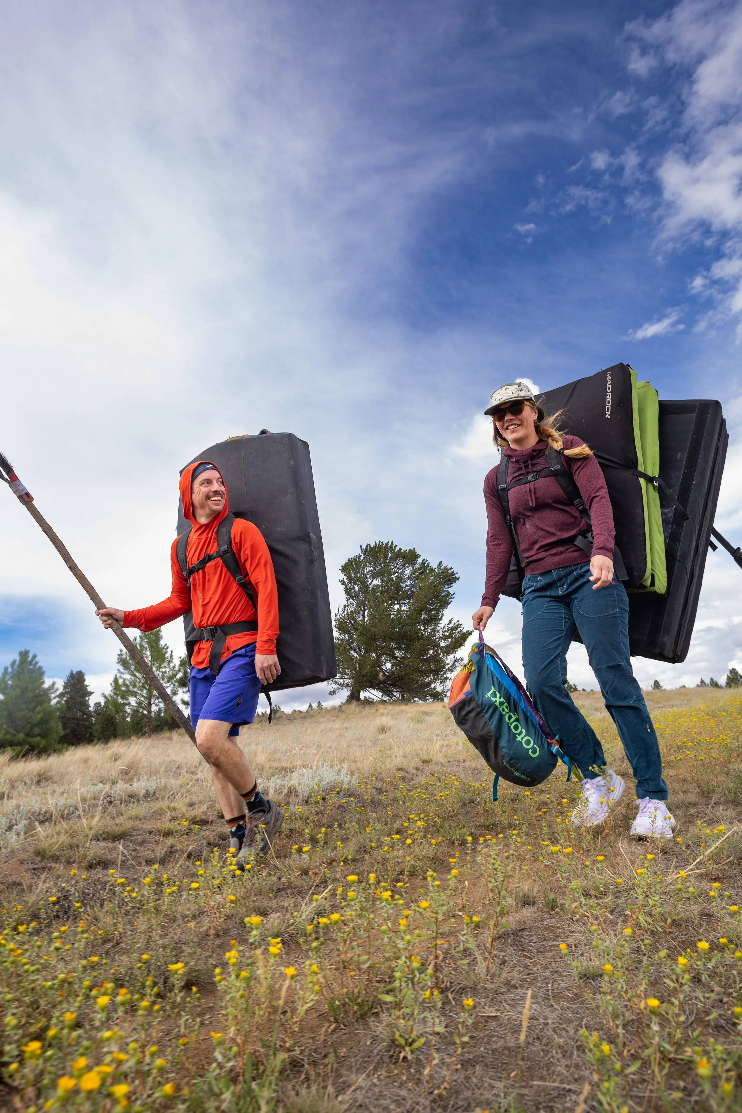 A man and woman hiking outdoors with large backpacks, smiling and enjoying the walk on a grassy hill under a blue sky with some clouds.