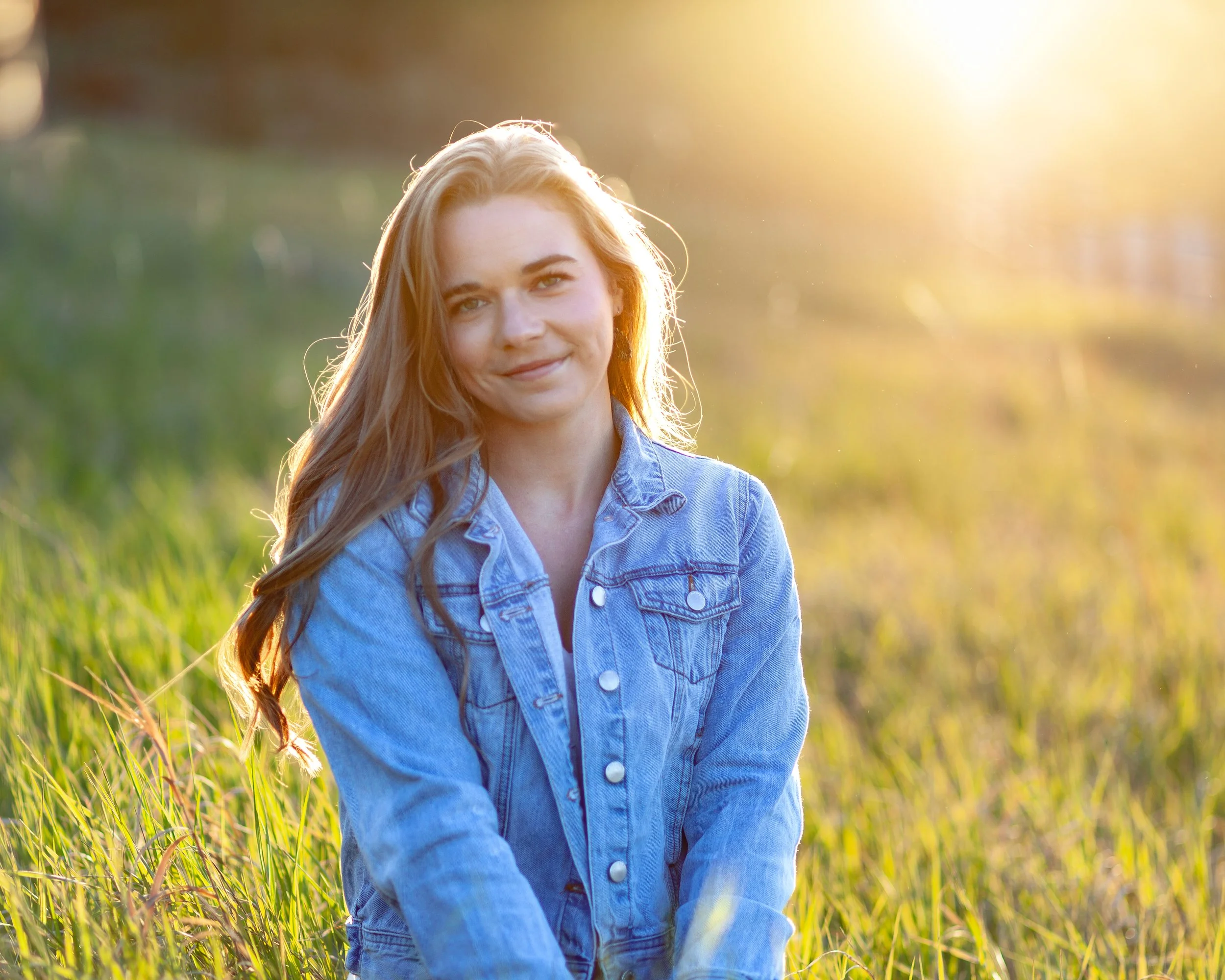 Young woman with long hair smiling in a grassy field during sunset, wearing a denim jacket.