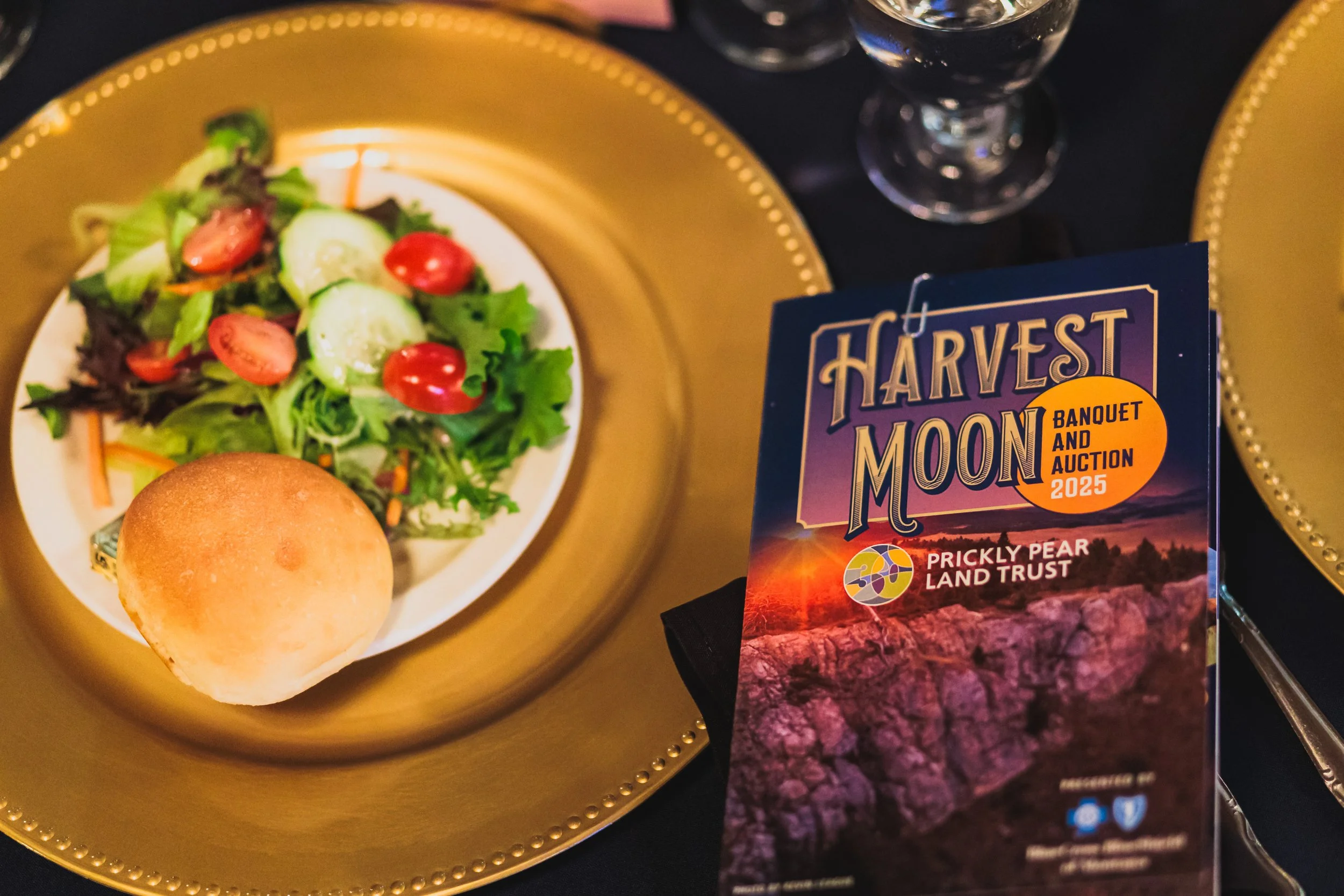 A plate with a small green salad containing cherry tomatoes, cucumber slices, and leafy greens, alongside a dinner roll on a gold-colored tray; next to a event program for 'Harvest Moon Banquet and Auction 2025' organized by Prickly Pear Land Trust, 