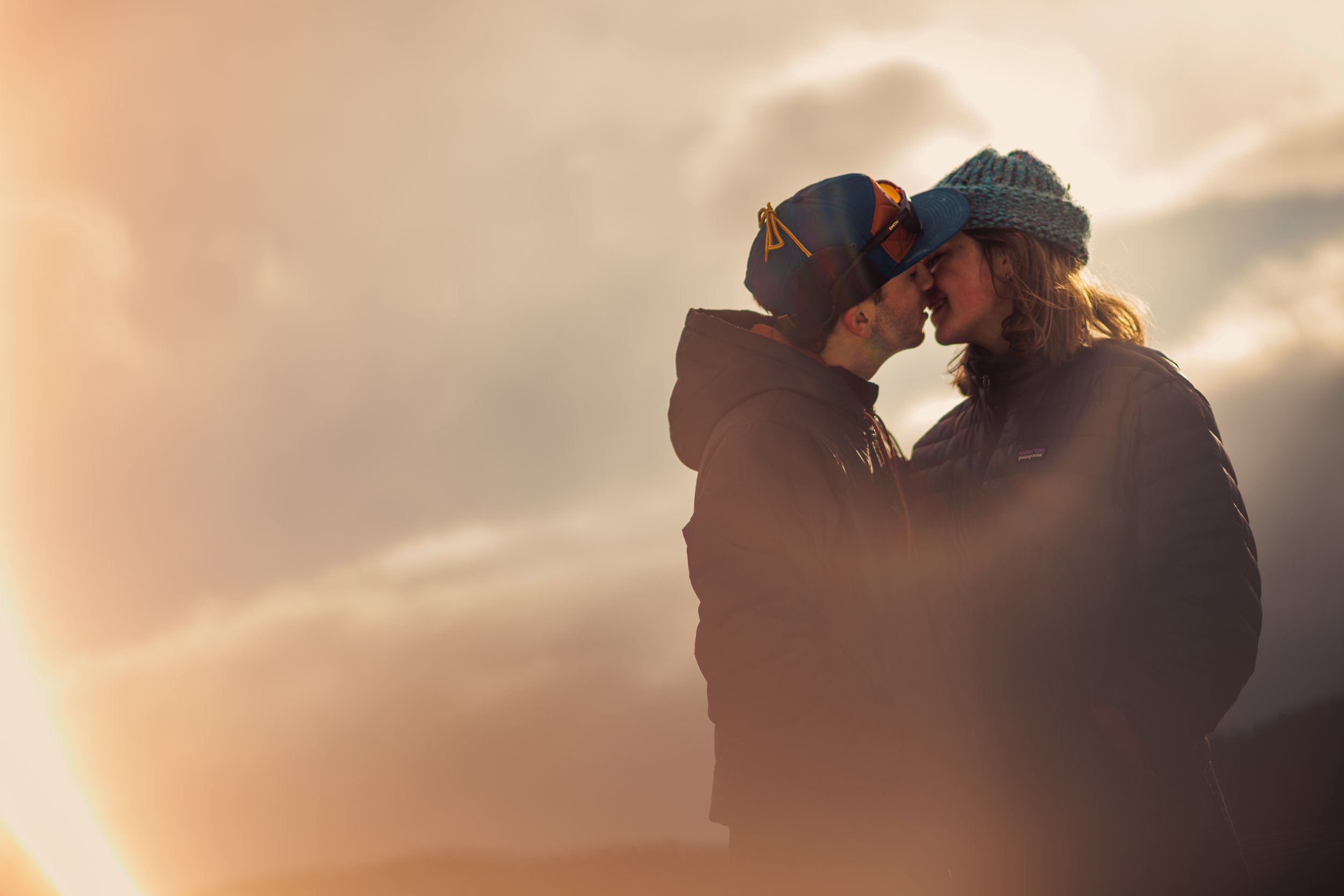 A couple sharing a kiss outdoors at sunset, with a cloudy sky in the background, wearing outdoor jackets and hats.