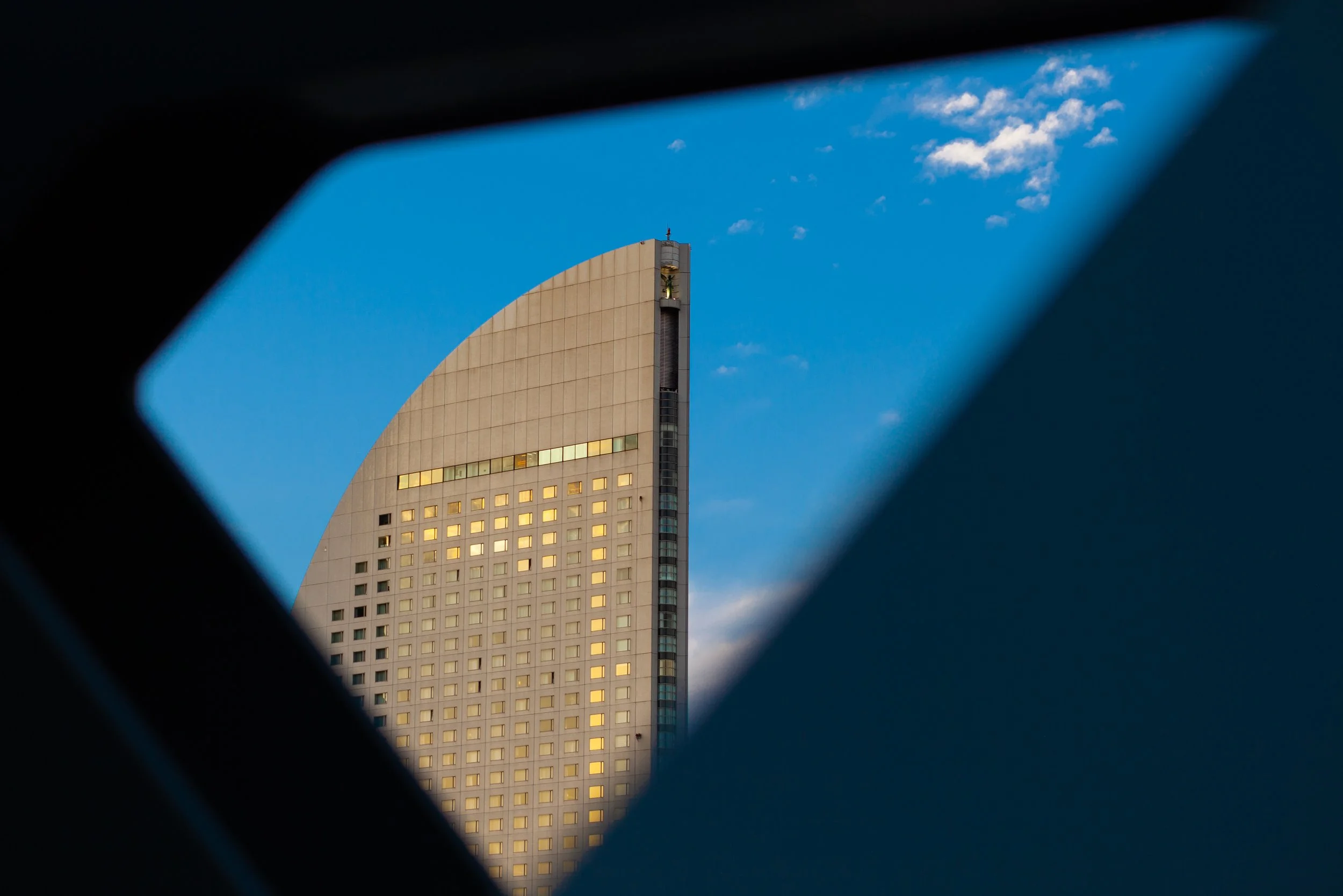 View of a modern high-rise building through a geometric window frame, with a clear blue sky and a few clouds in the background.