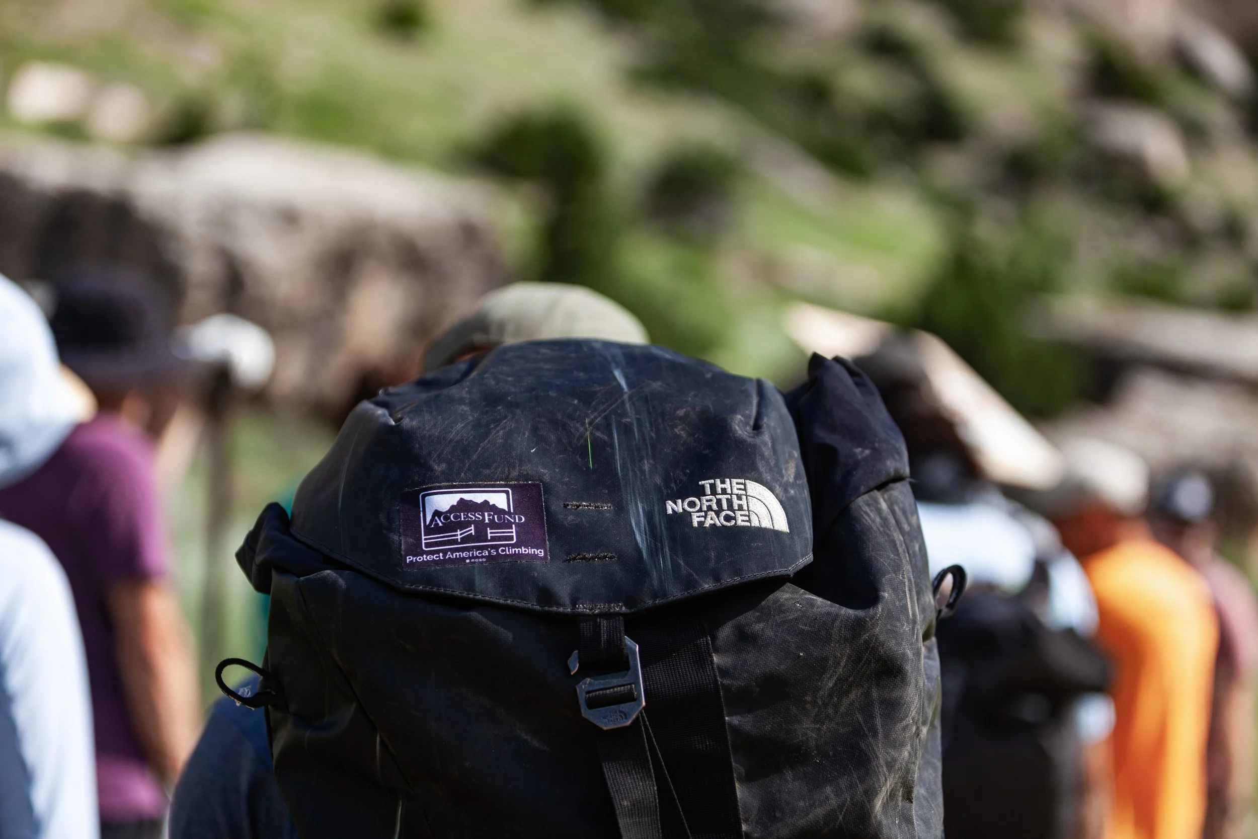 Backpack with The North Face logo and Access Fund sticker attached, outdoors with people in the background.