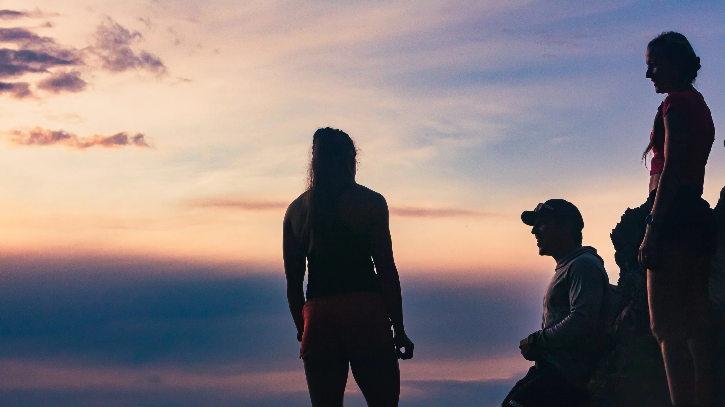 Silhouettes of three women and one man during sunset, with one woman standing and two women sitting on rocks, and a man sitting, all on a rocky area.