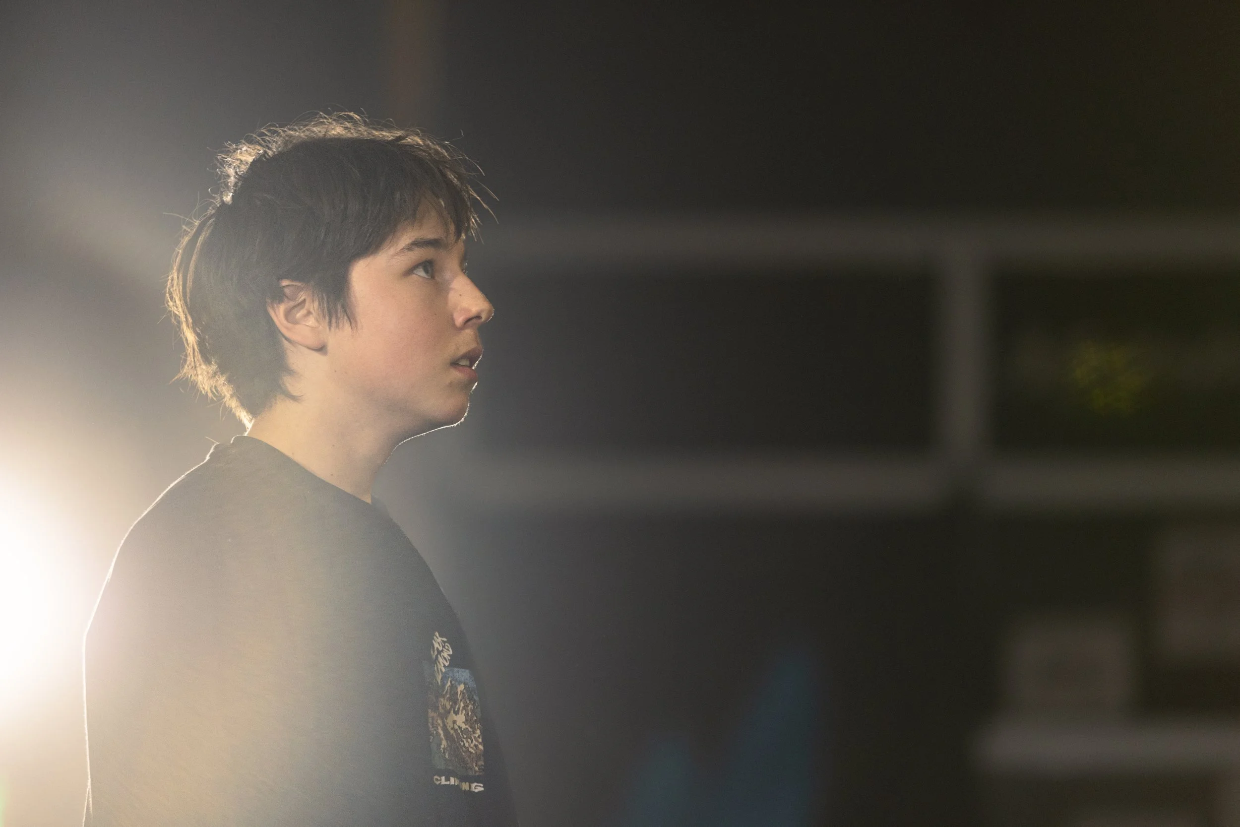 Side profile of a young male with dark hair, looking to the left, with a bright light behind him on the left side of the image in a dimly lit environment.