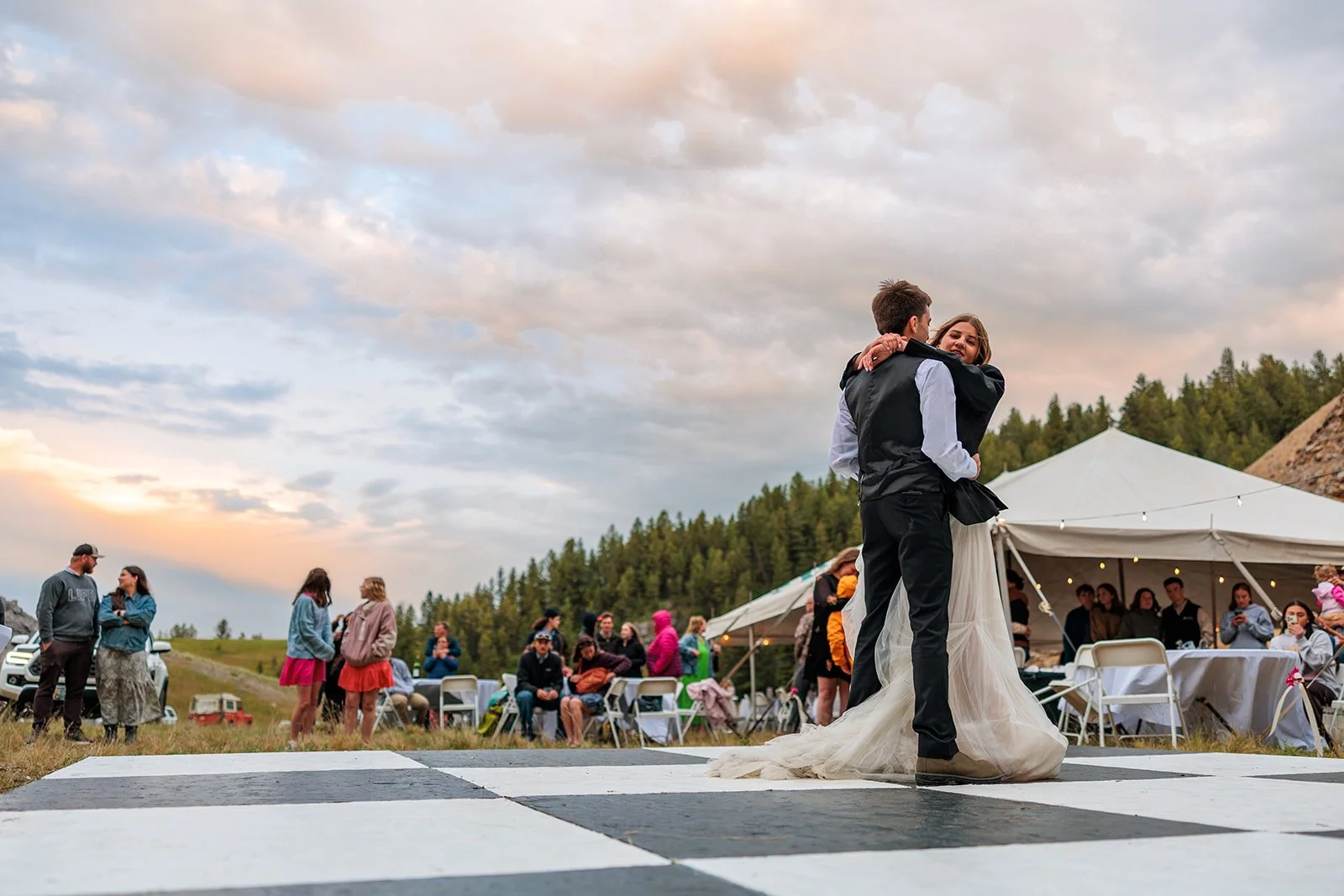 A couple dancing on a checkered dance floor outdoors during sunset at a wedding reception, with guests and a large tent in the background.
