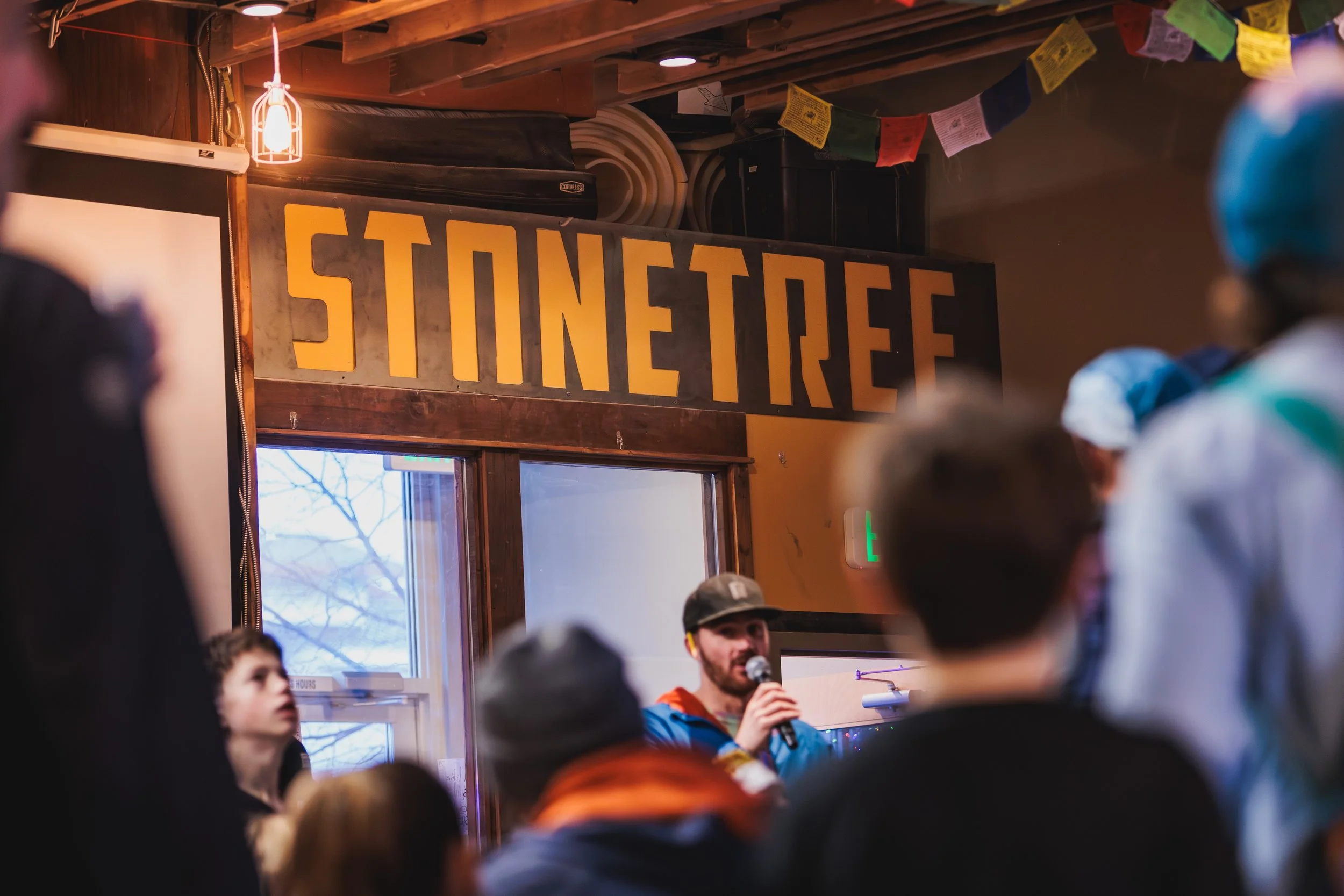 Indoor gathering with people listening to a speaker, a large sign reading 'STINETREE' hangs on the wall, and colorful flags decorate the ceiling.